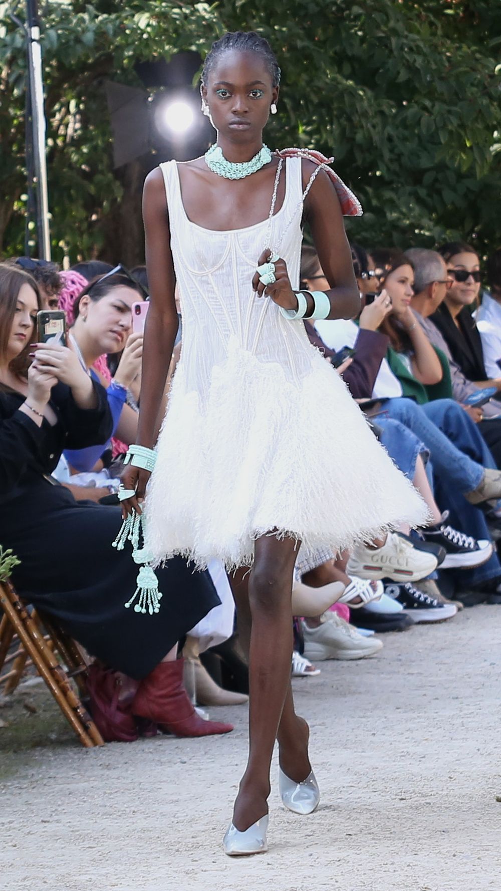 White feathered mini dress with ruffles shown on outdoor catwalk with audience seated along path.