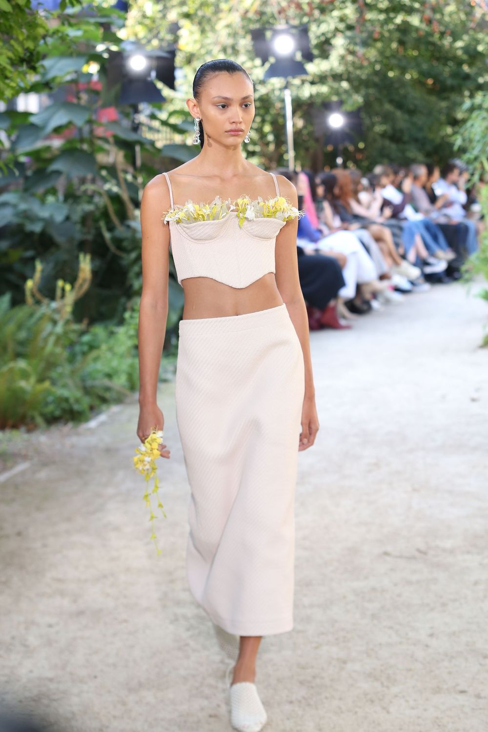 Model in pale pink dress with ruffle details walks outdoor fashion show runway.