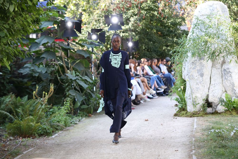 Outdoor fashion show with audience seated along tree-lined gravel runway path.