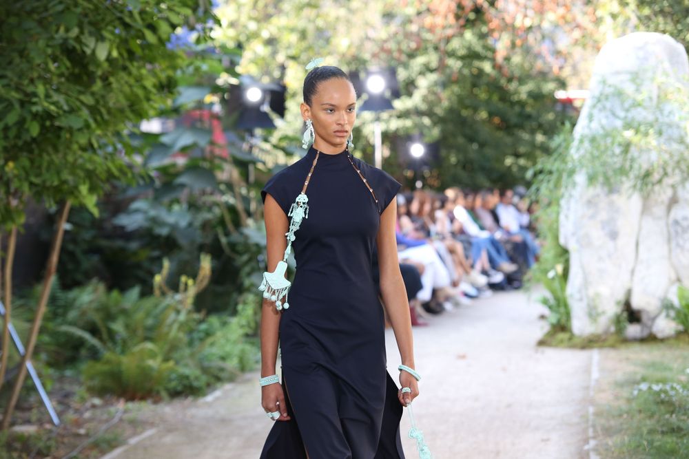 Model wearing simple black outfit walking down garden path at fashion show.