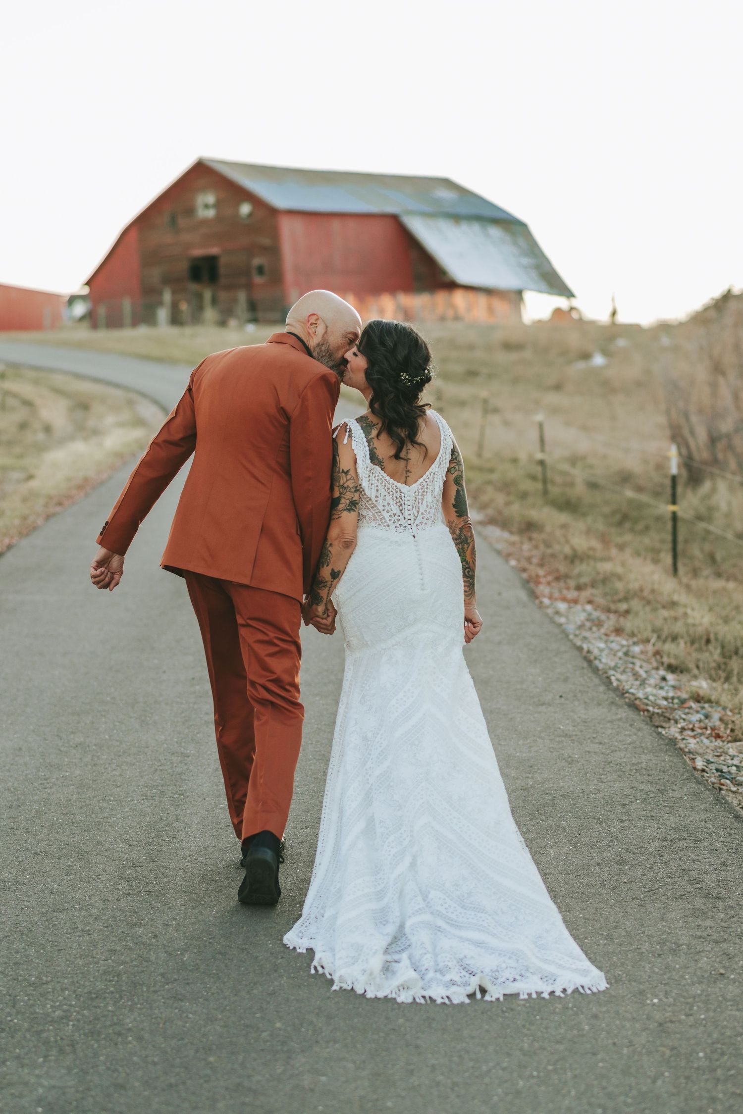 A bride in a lace dress and groom in orange suit pose together at sunset with a farmhouse in the background.
