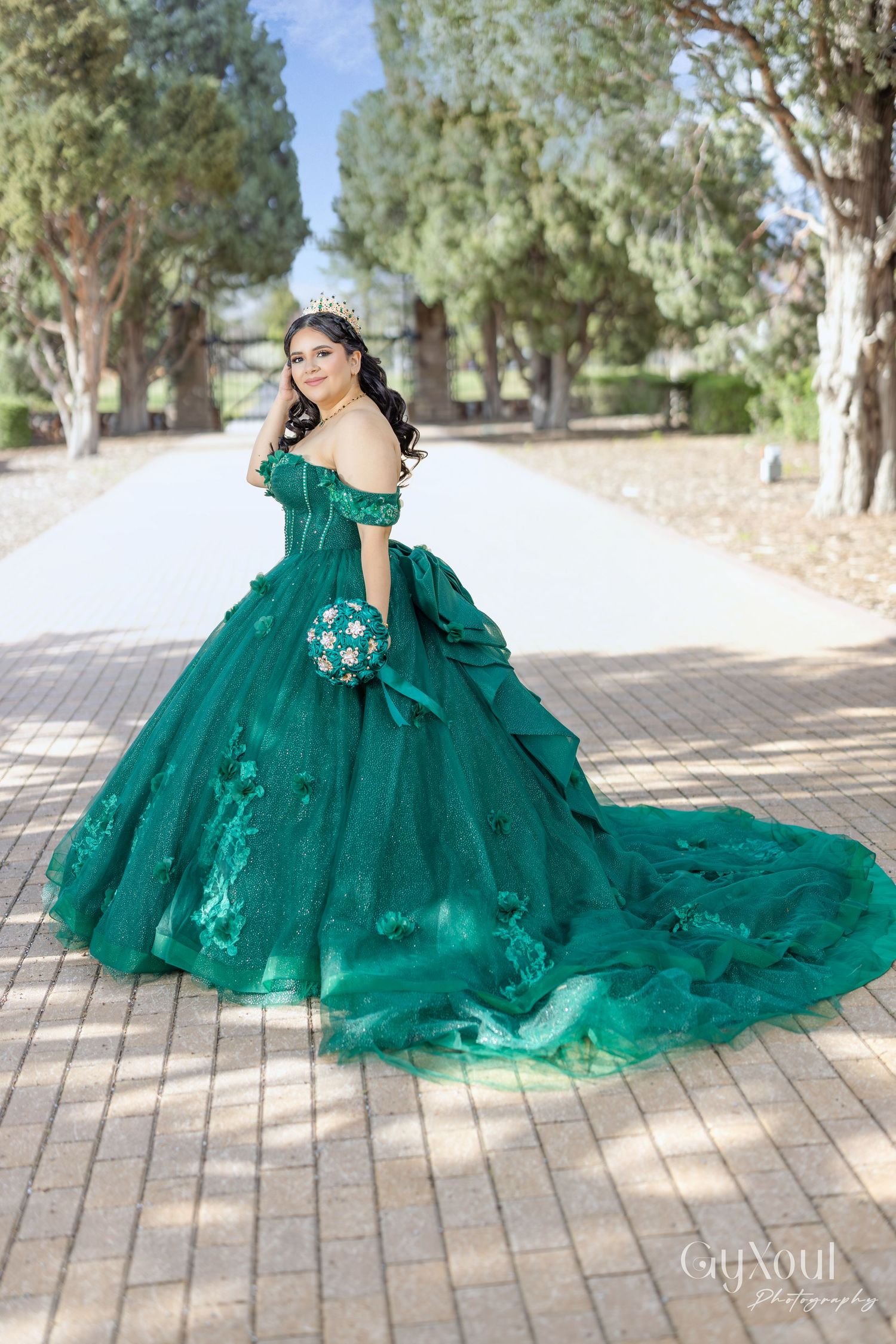 A stunning emerald green ball gown with tulle layers sweeps across a sunlit outdoor pathway.