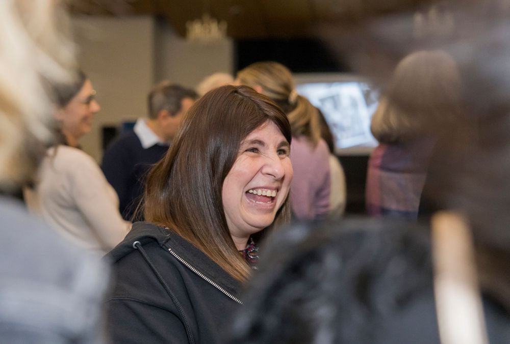 A person smiles warmly during an indoor gathering, wearing a black jacket.