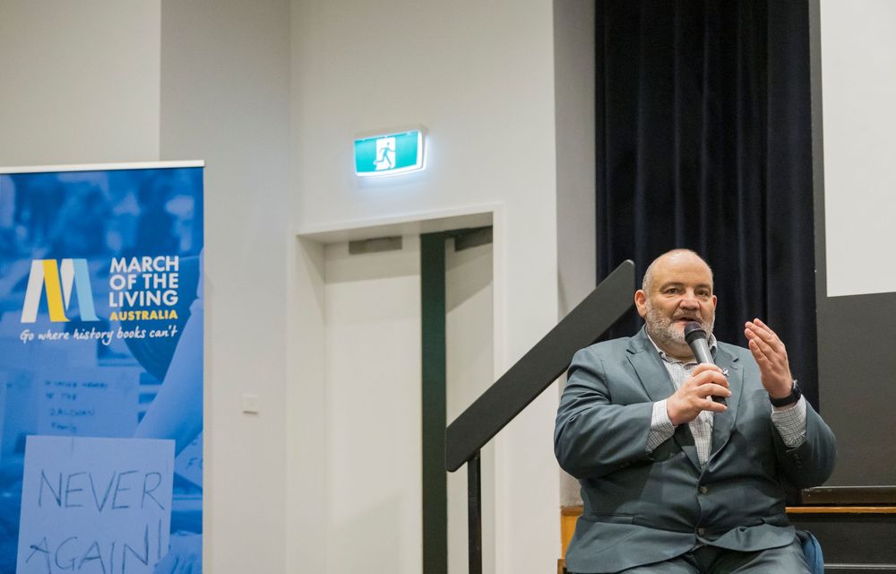 A speaker in a dark suit gestures while holding a microphone during a presentation with a blue banner visible on the wall.