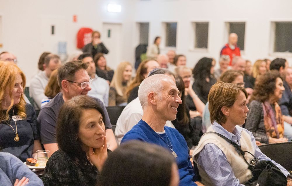 A diverse audience smiles and laughs while seated in rows at an indoor event or presentation.