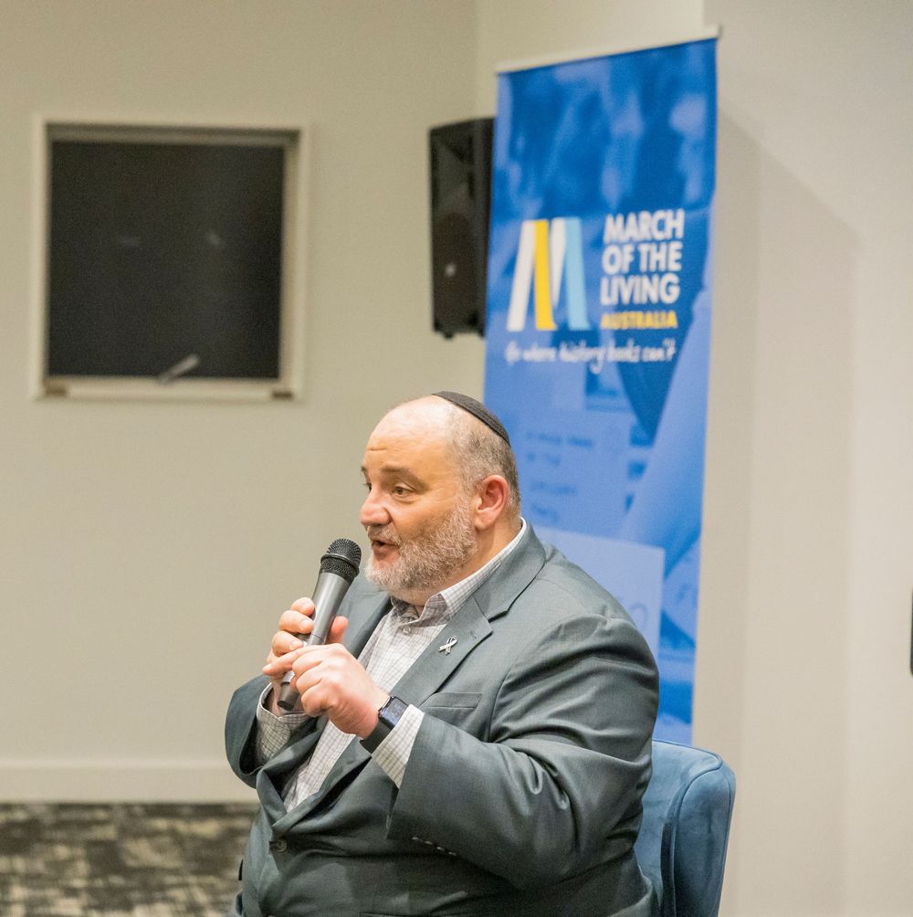 A person in a gray suit speaks into a microphone while seated at a panel discussion event.