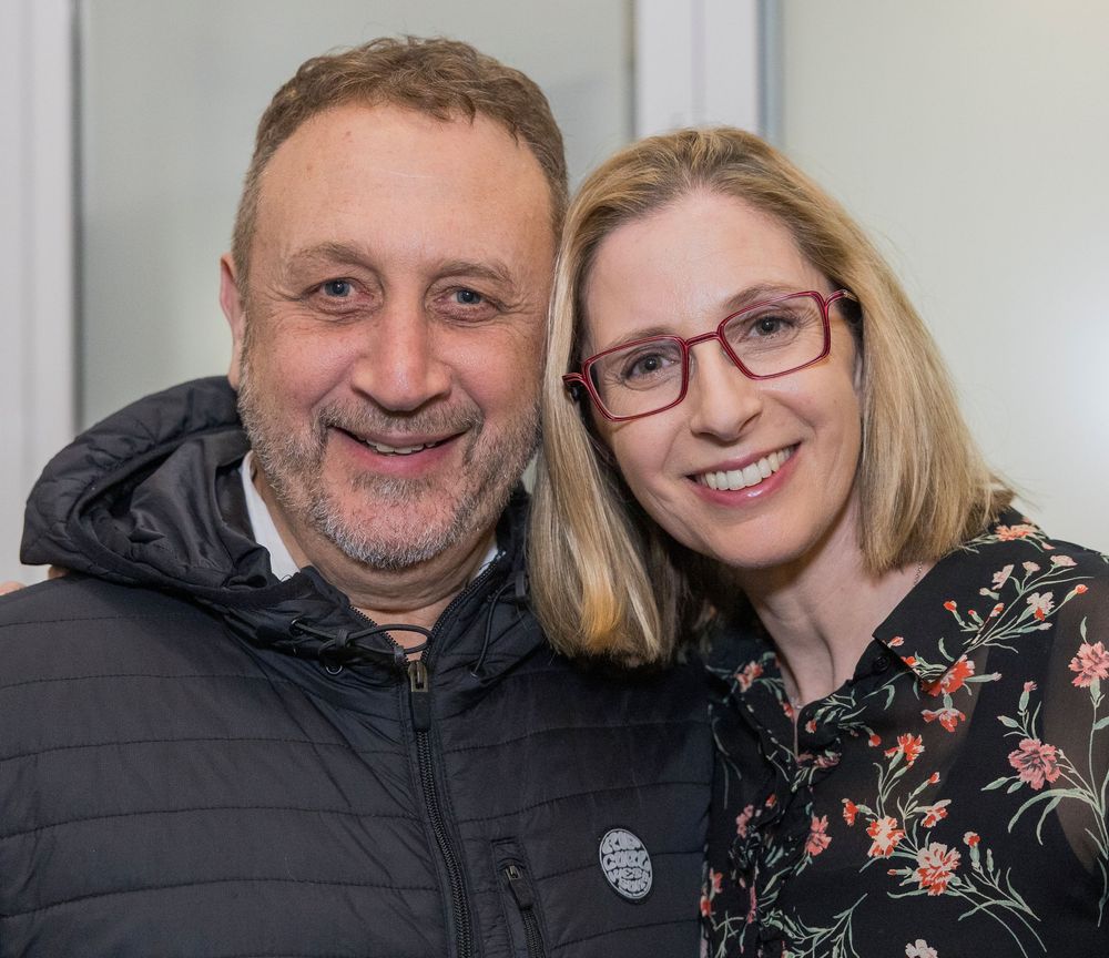 A smiling couple poses together, with one wearing a black puffy jacket and the other in glasses and a floral print top.