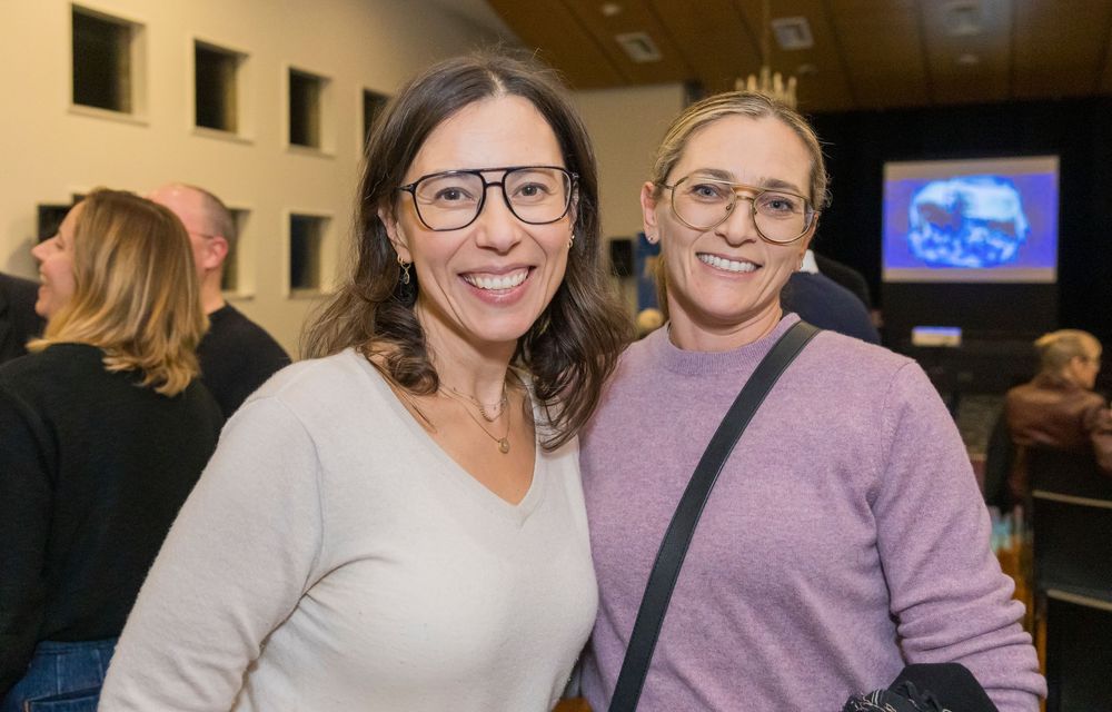 Three individuals wearing similar casual attire and glasses smile together at an indoor event.