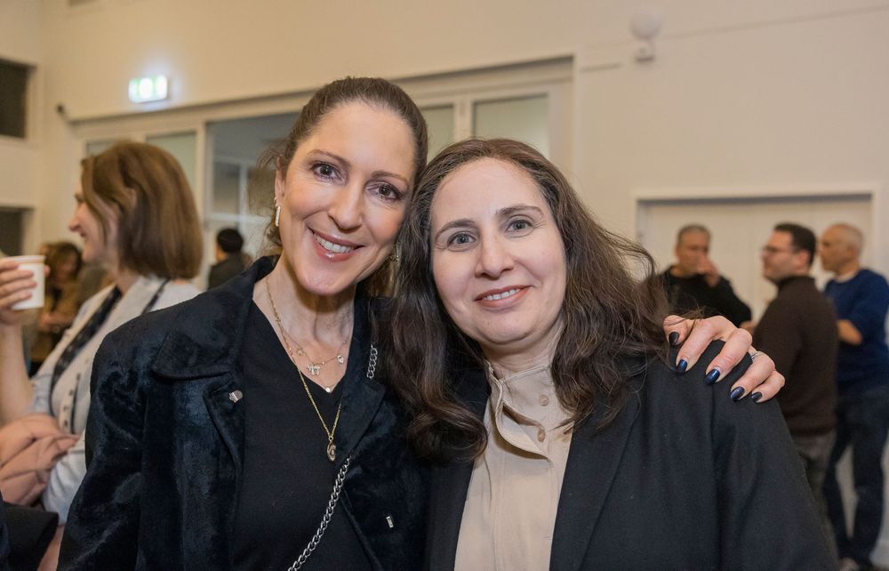 Two women smile together at a social gathering with people visible in the background.