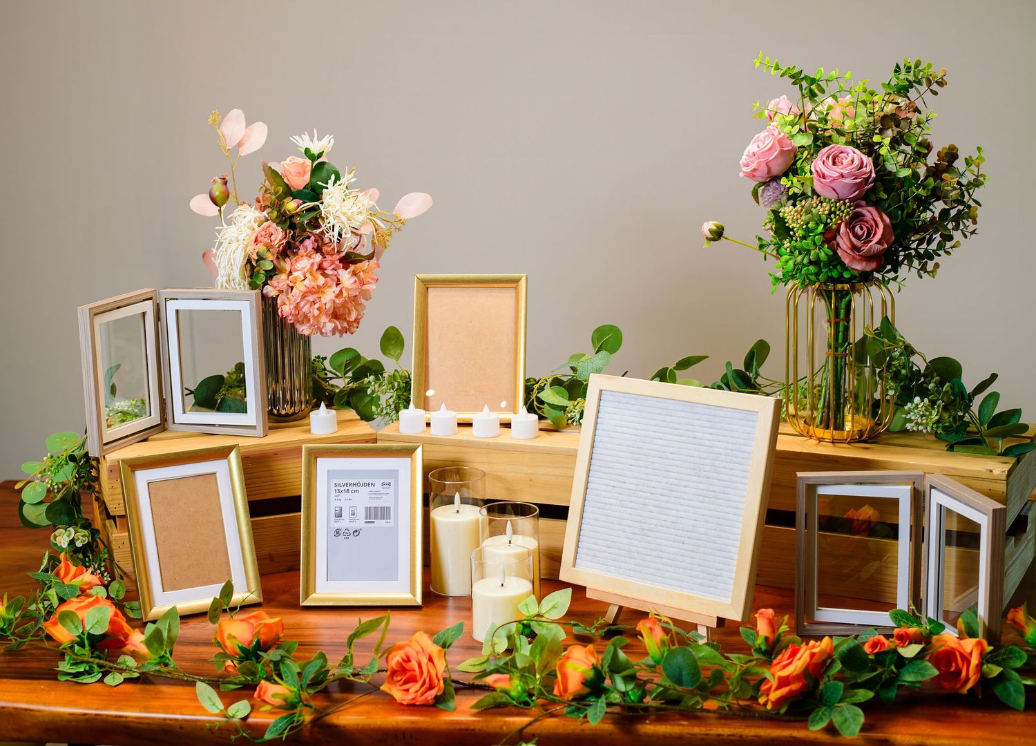 Elegant wedding reception display with empty picture frames, coral roses and greenery arrangements on wooden table.