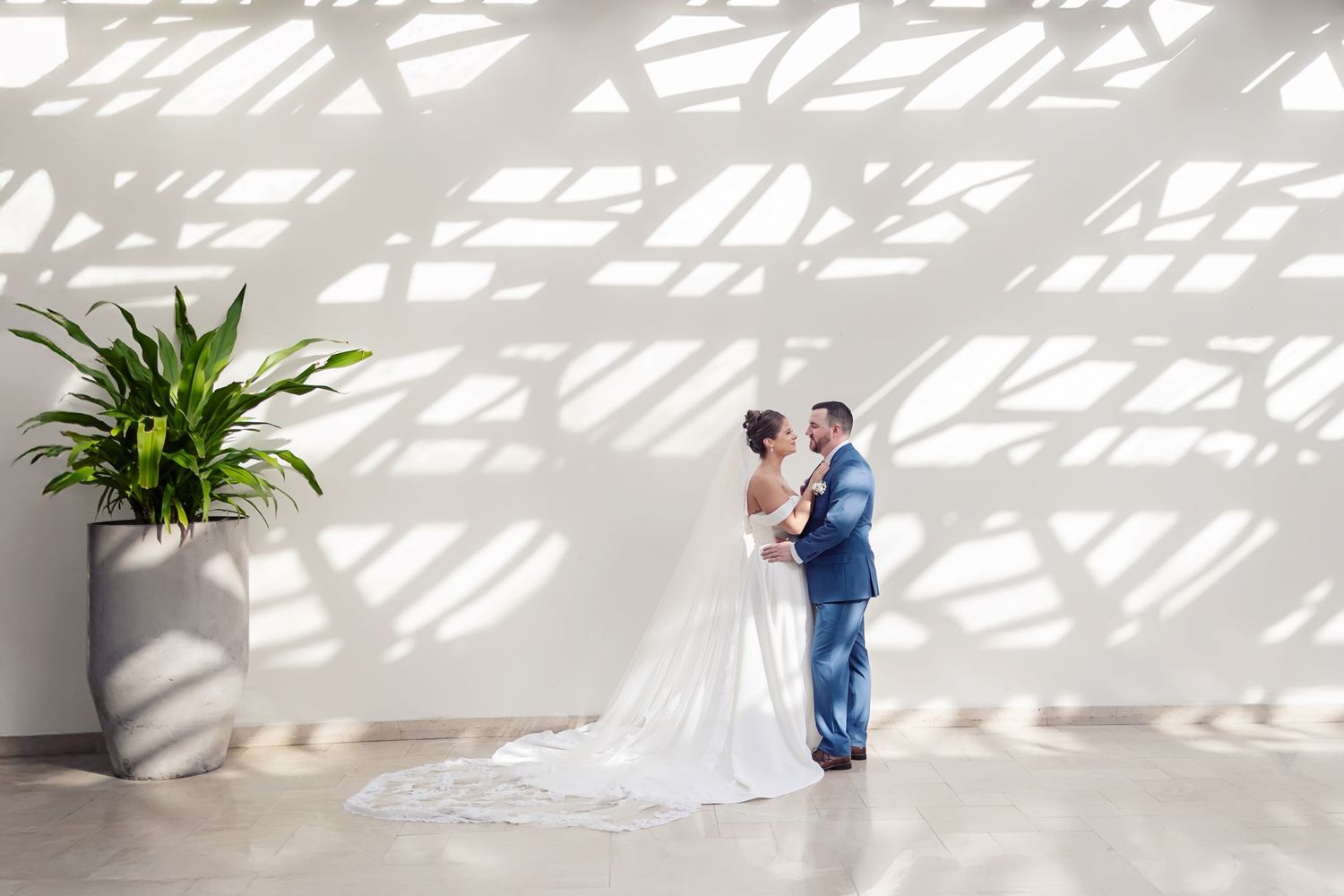 A couple shares an intimate moment on their wedding day against a white wall with dramatic shadow patterns and a potted plant.