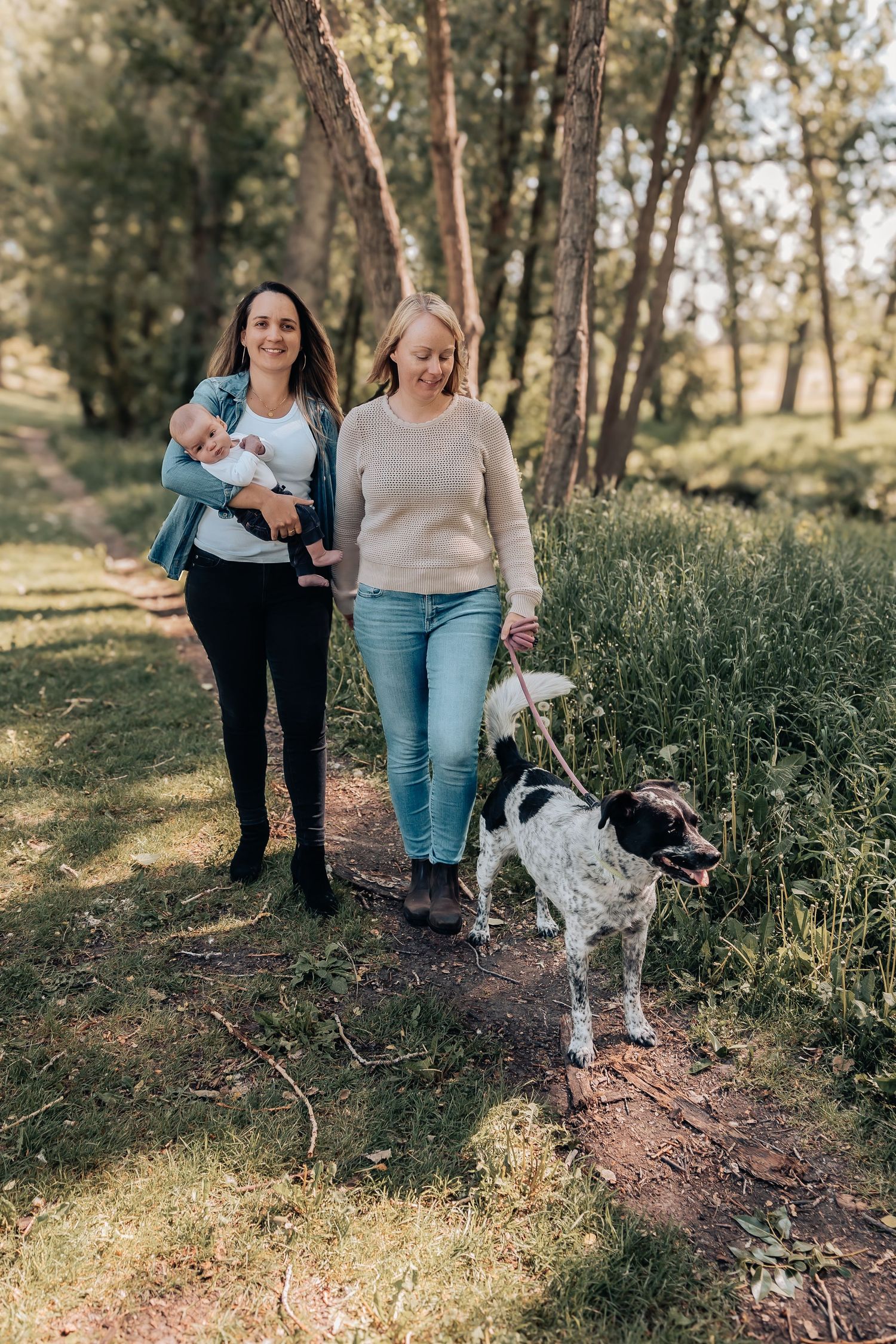 People walking on a forest trail with a small white dog amid tall grass and trees on a sunny afternoon in Calgary, Alberta.