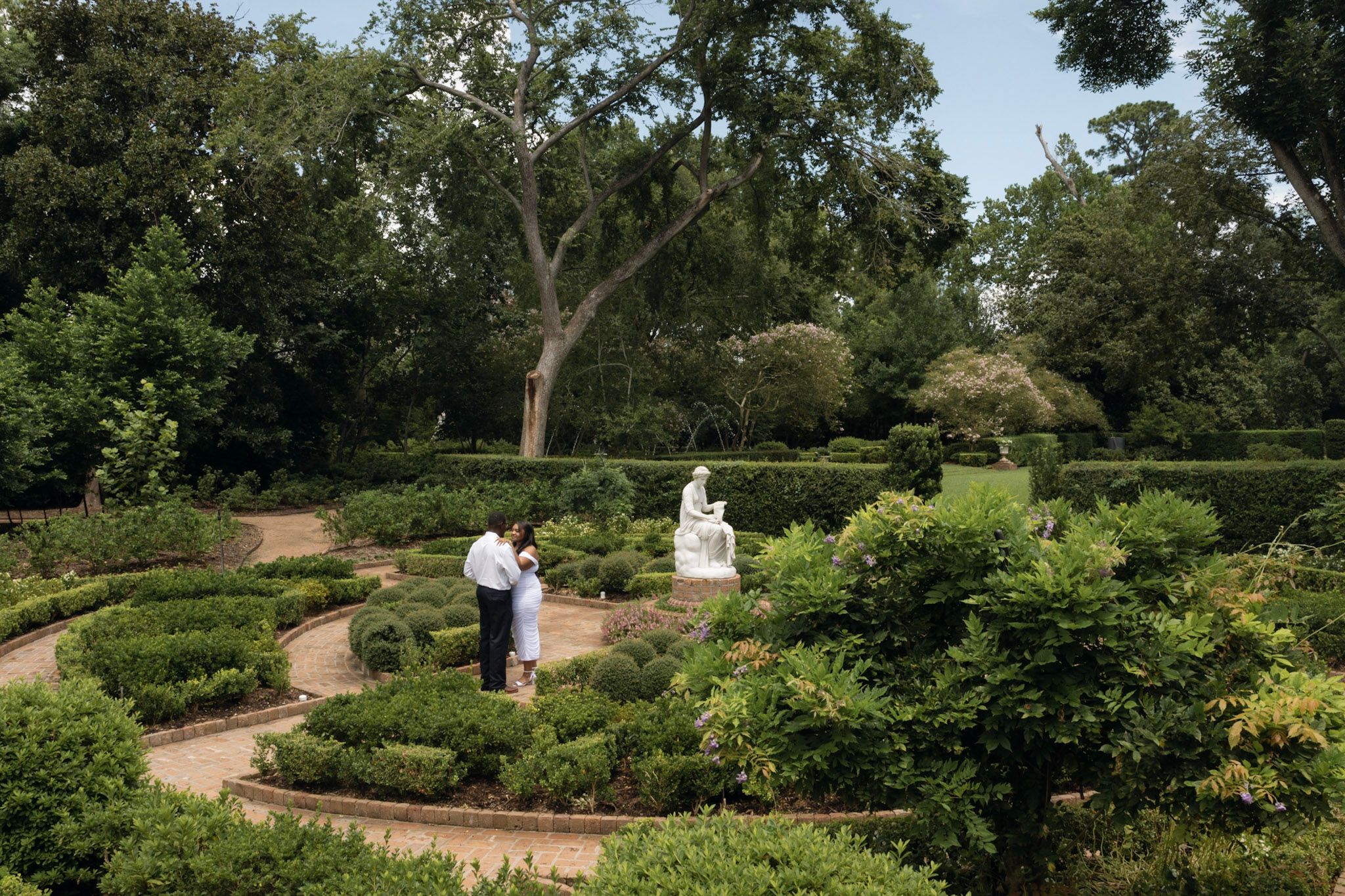 Two people in white clothing standing on a winding garden path surrounded by greenery.