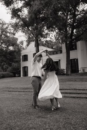 A couple dances joyfully on a lawn in black and white, with a white manor house and trees in the background.