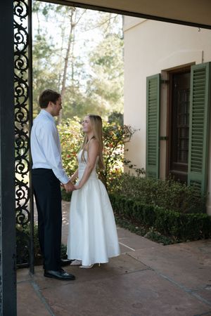 Bride and groom holding hands in white dress and suit by ornate iron gate with green shuttered building.