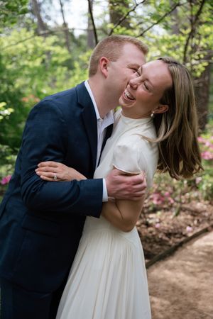 A joyful couple embraces outdoors, woman in white dress laughing as man in navy suit nuzzles her cheek.
