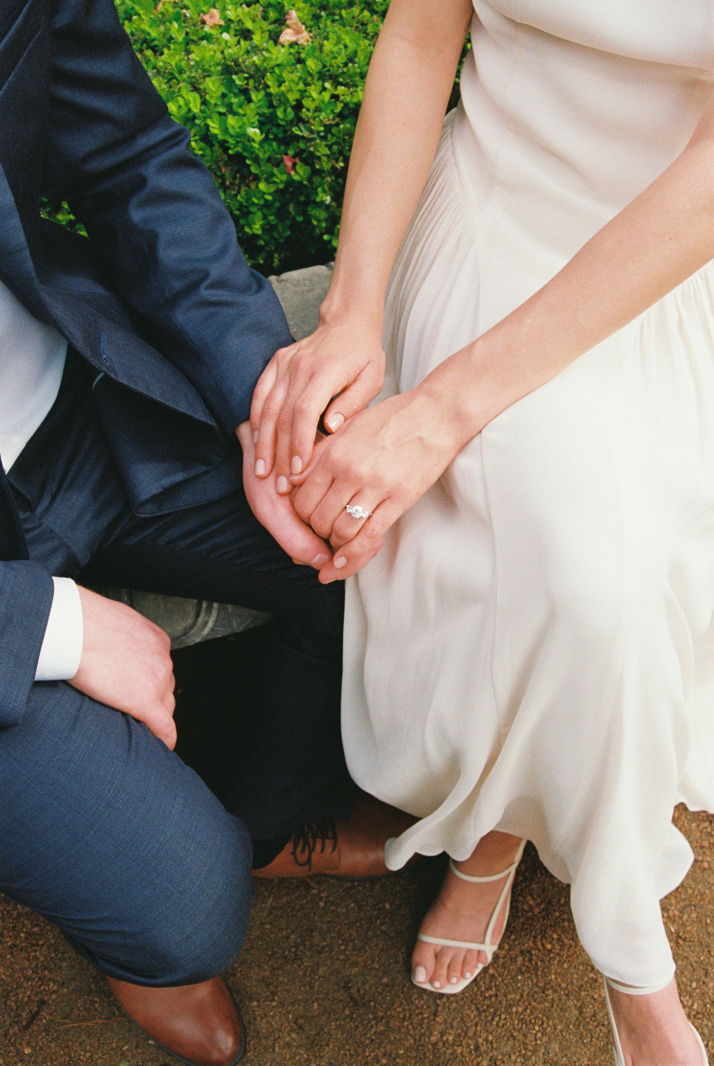 Bride and groom holding hands, showcasing wedding ring, bride in white dress, groom in navy suit, seated outdoors.