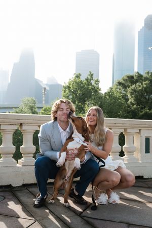 Couple sitting on stone balustrade with their brown and white dog licking the man's face, city skyline backdrop.