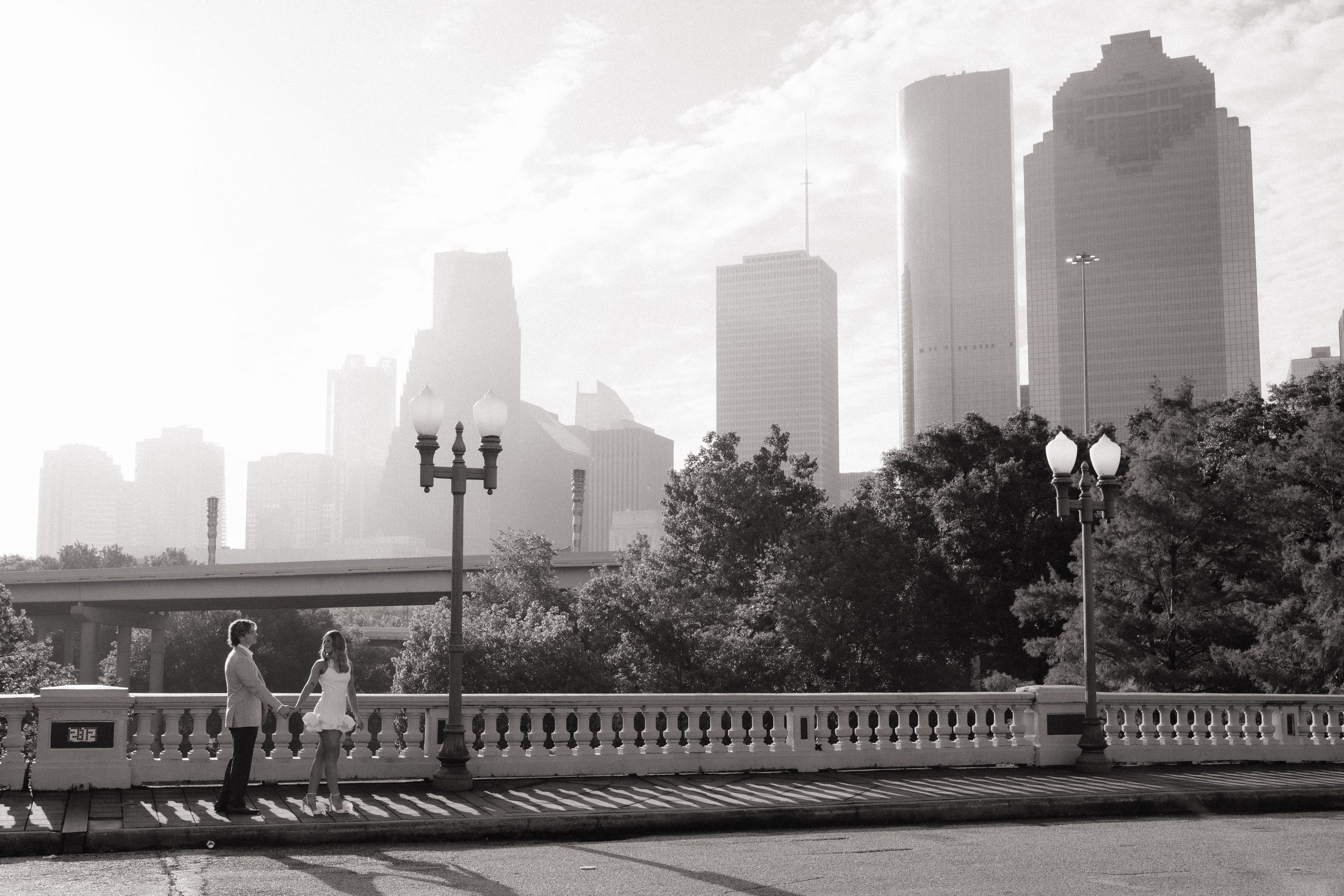 Black and white photo of two people on a bridge with Houston city skyline and trees in the background.