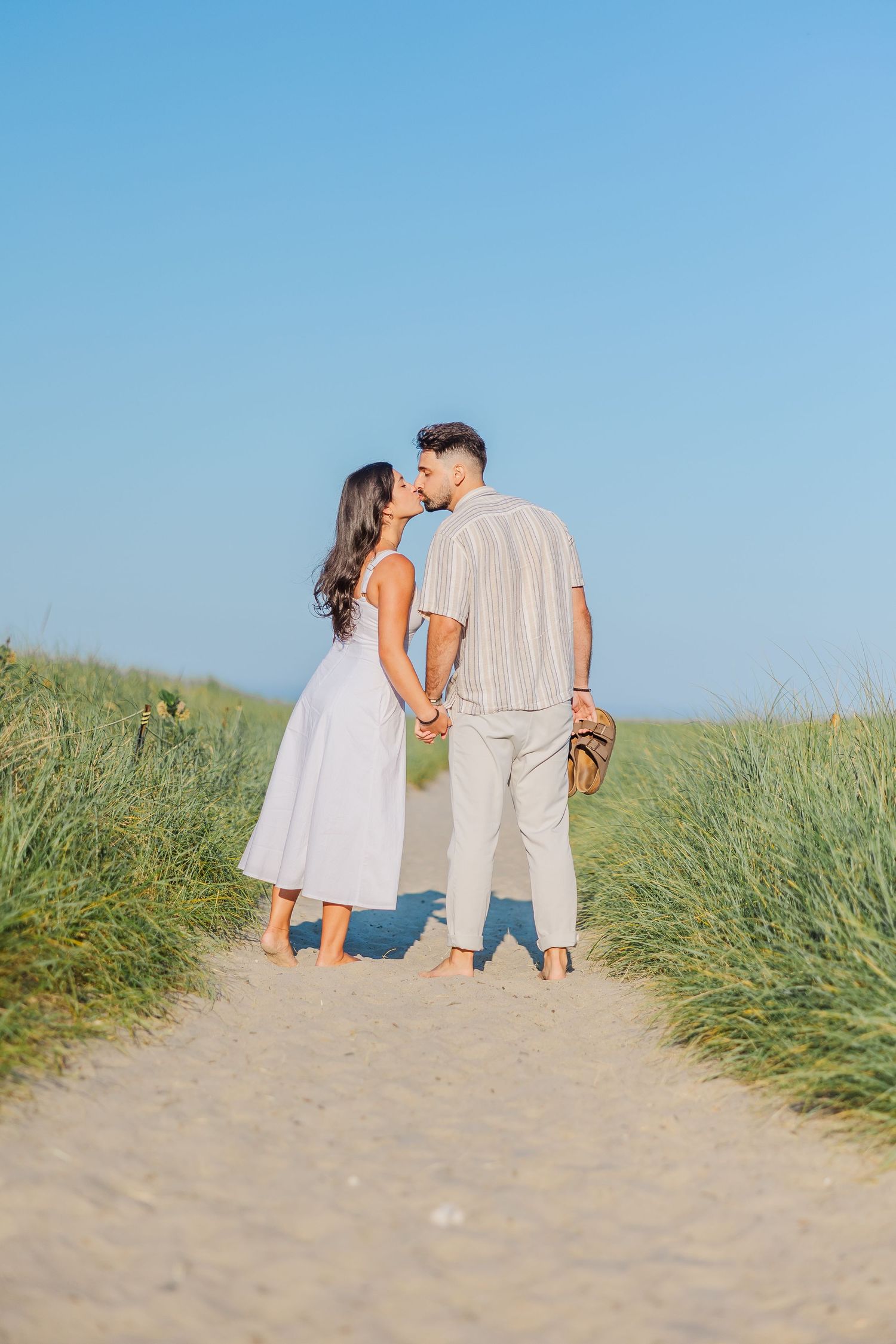 A couple in light summer attire share an intimate moment while standing on a sandy path between tall grass on a sunny day.