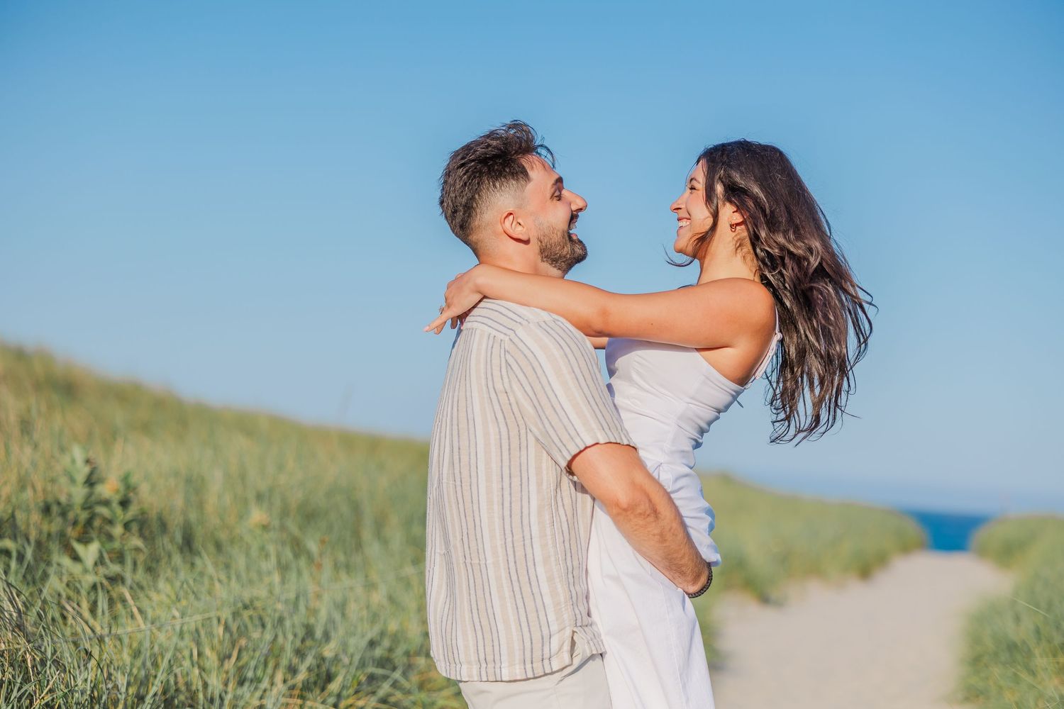 A couple embraces and smiles together during a seaside photoshoot in a grassy coastal setting.