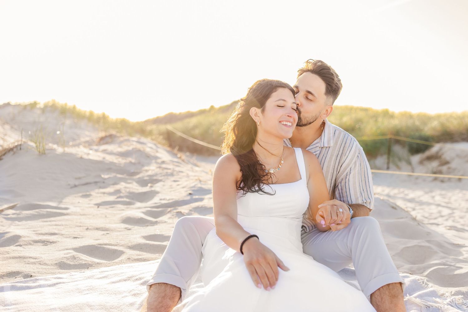 A romantic couple sitting together on a sandy beach at sunset in light colored clothing.