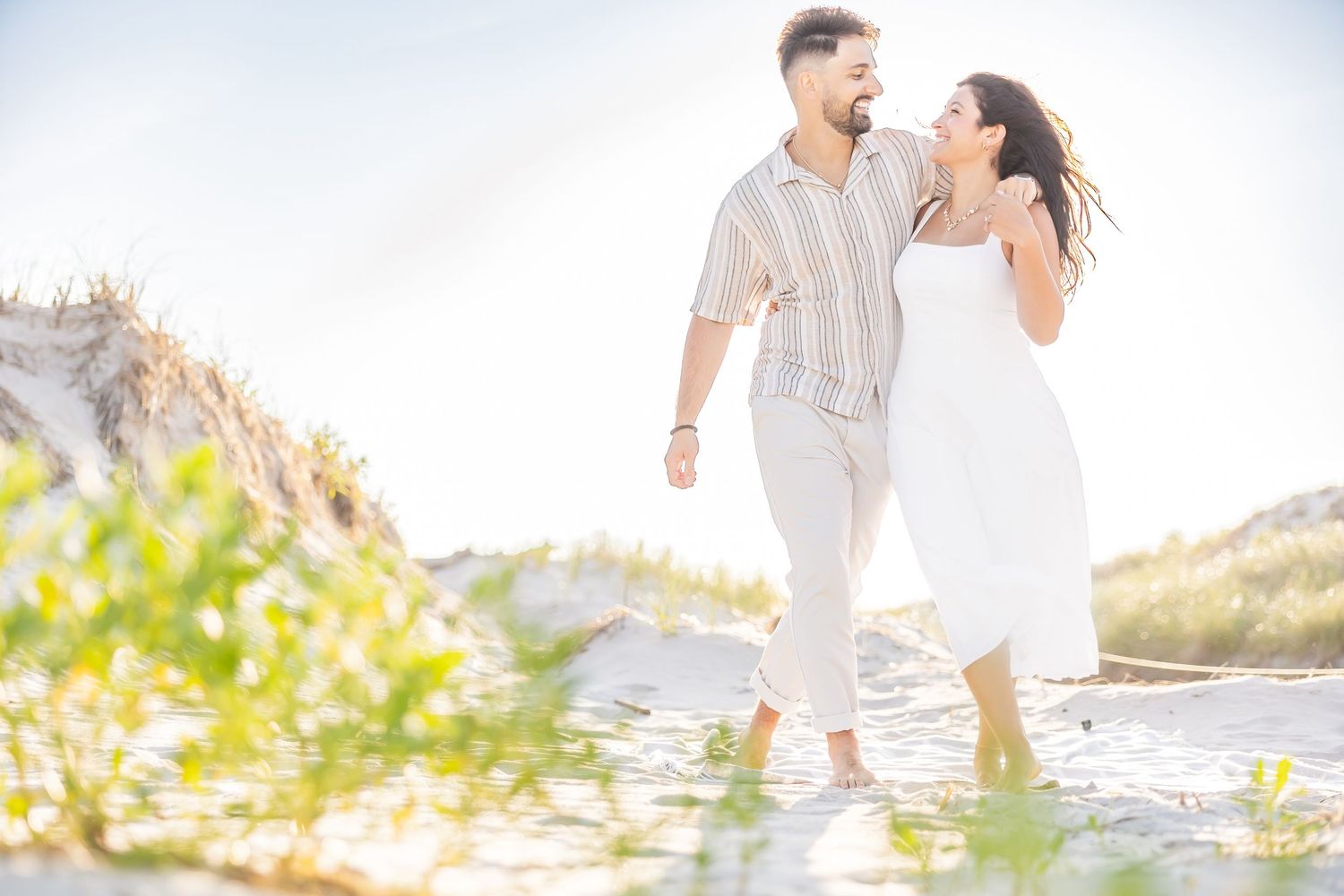 A romantic couple walks together in shallow water along a sunny beach with mountains in the background.