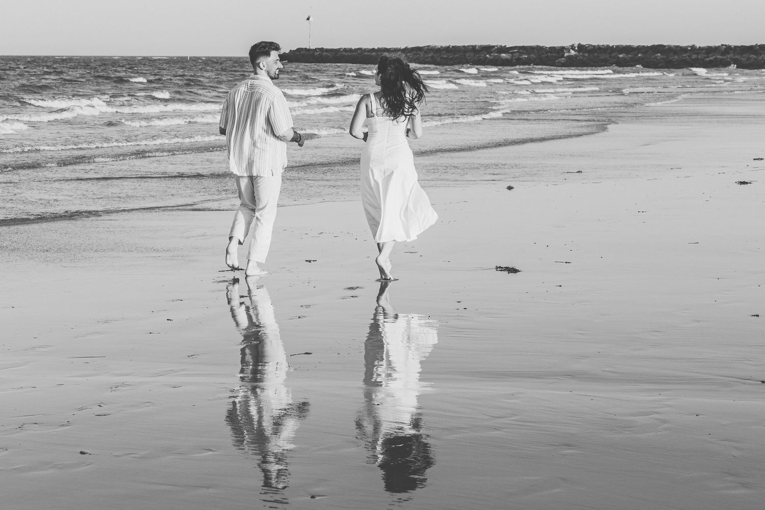 A couple in white clothing holds hands while walking along a beach shore with their reflections in the wet sand.