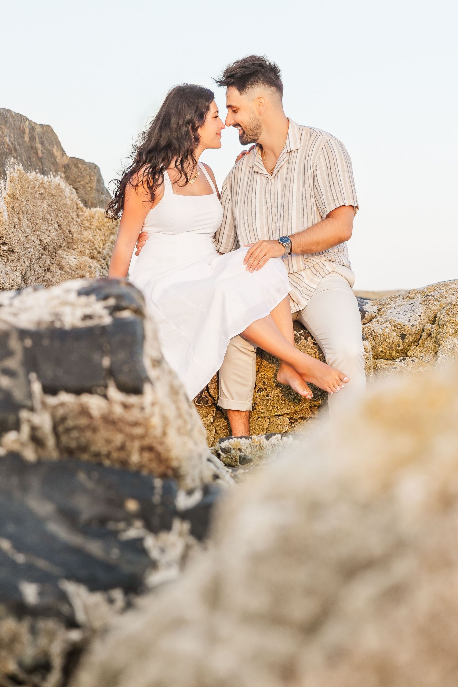 A romantic couple shares intimate moments on a rocky beach during a sunset photoshoot.