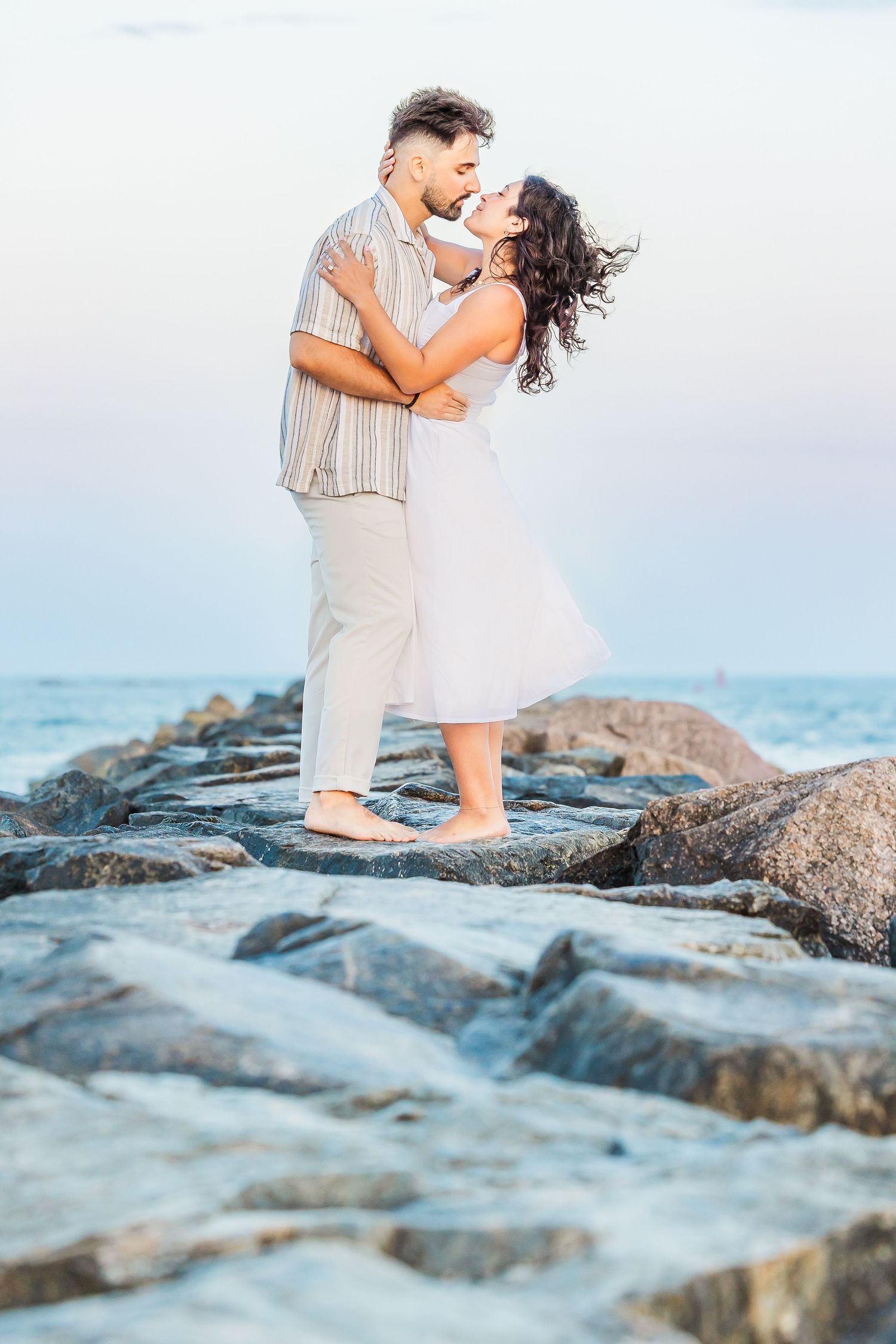 A couple embraces romantically on rocky coastal shores with ocean waves and soft blue sky in the background.