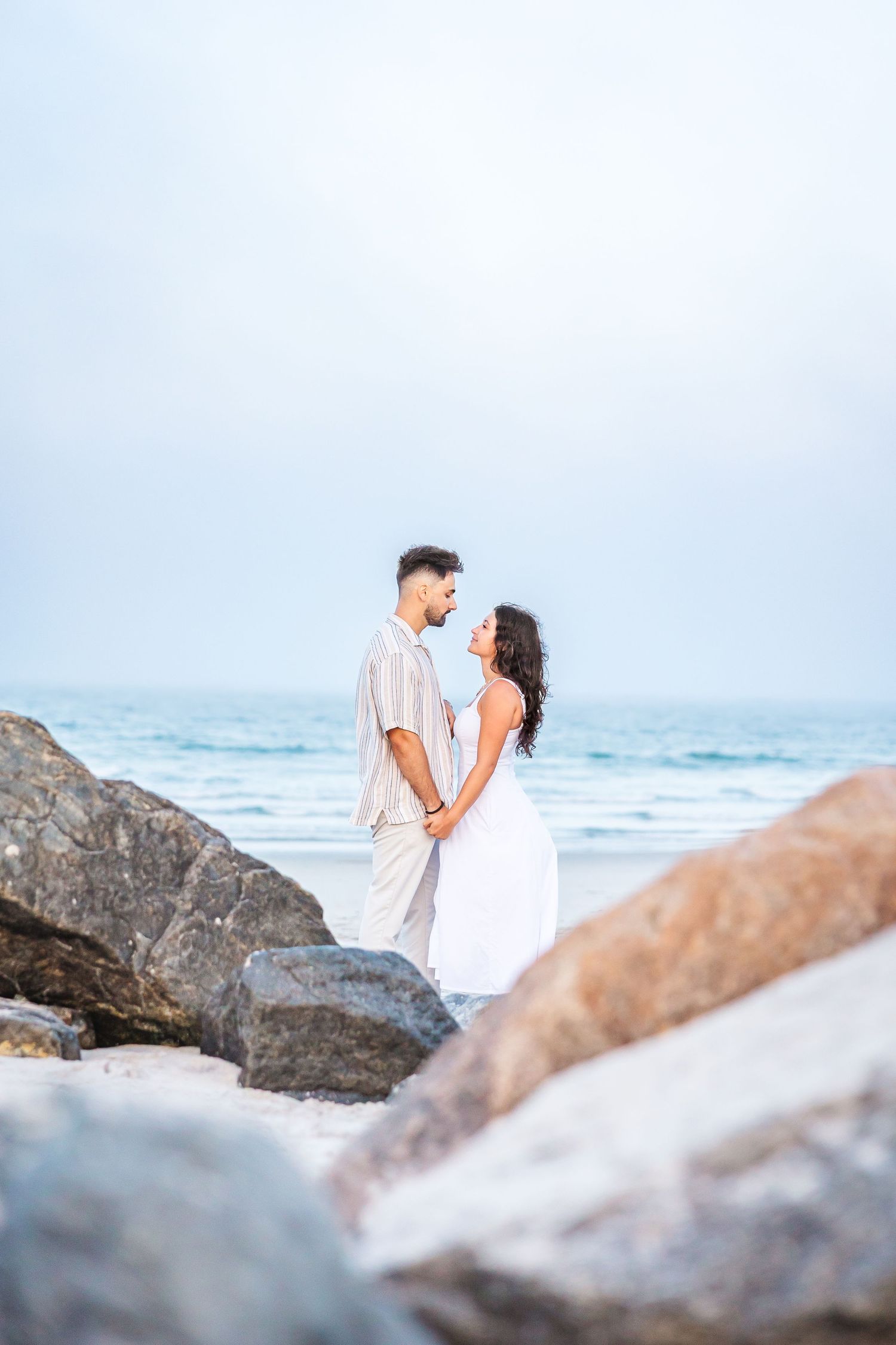 A couple in white outfits embraces on a rocky beach while waves crash in the background.