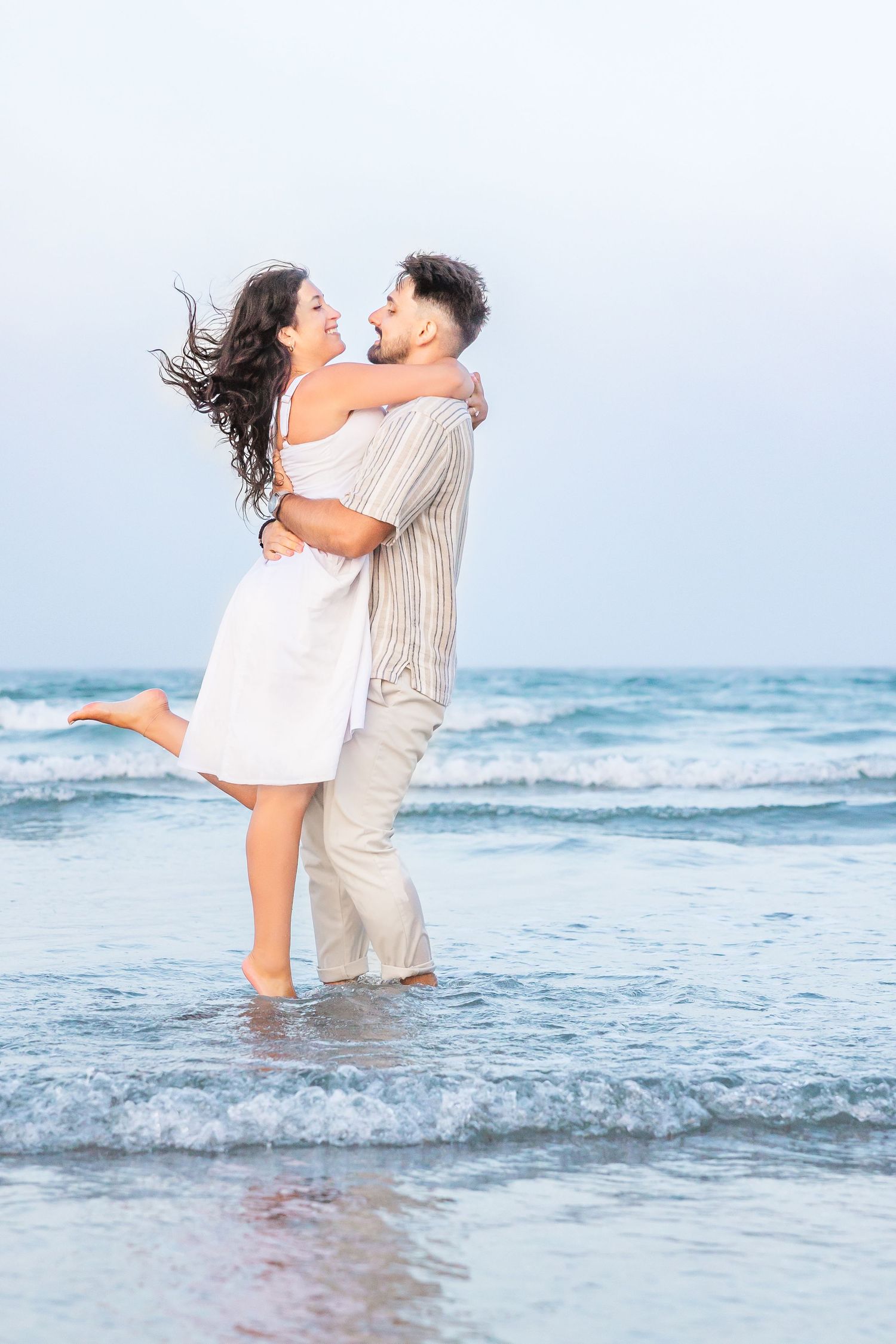 A couple embraces joyfully at the edge of ocean waves on a misty beach, their clothing flowing in the sea breeze.