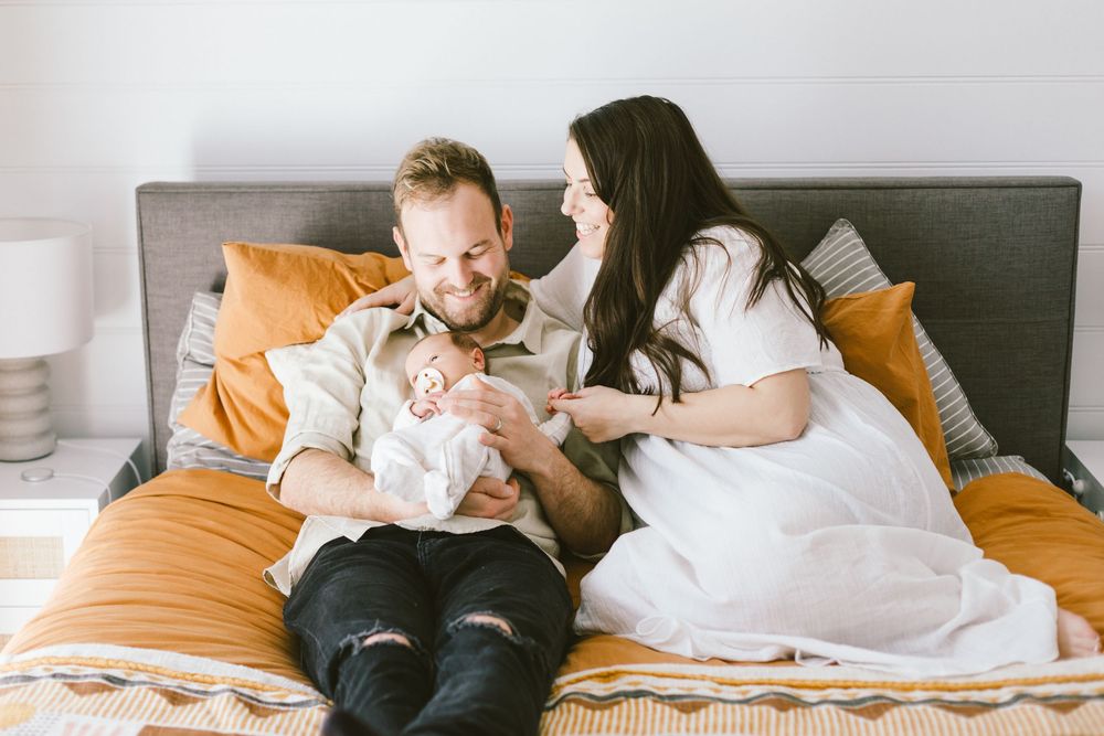 Family relaxing on bed with newborn baby and orange pillows.