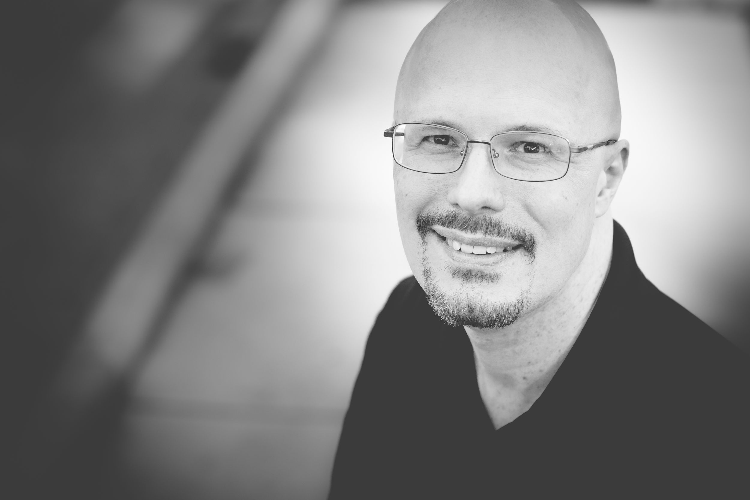 A black and white professional headshot with glasses against a gradient background.