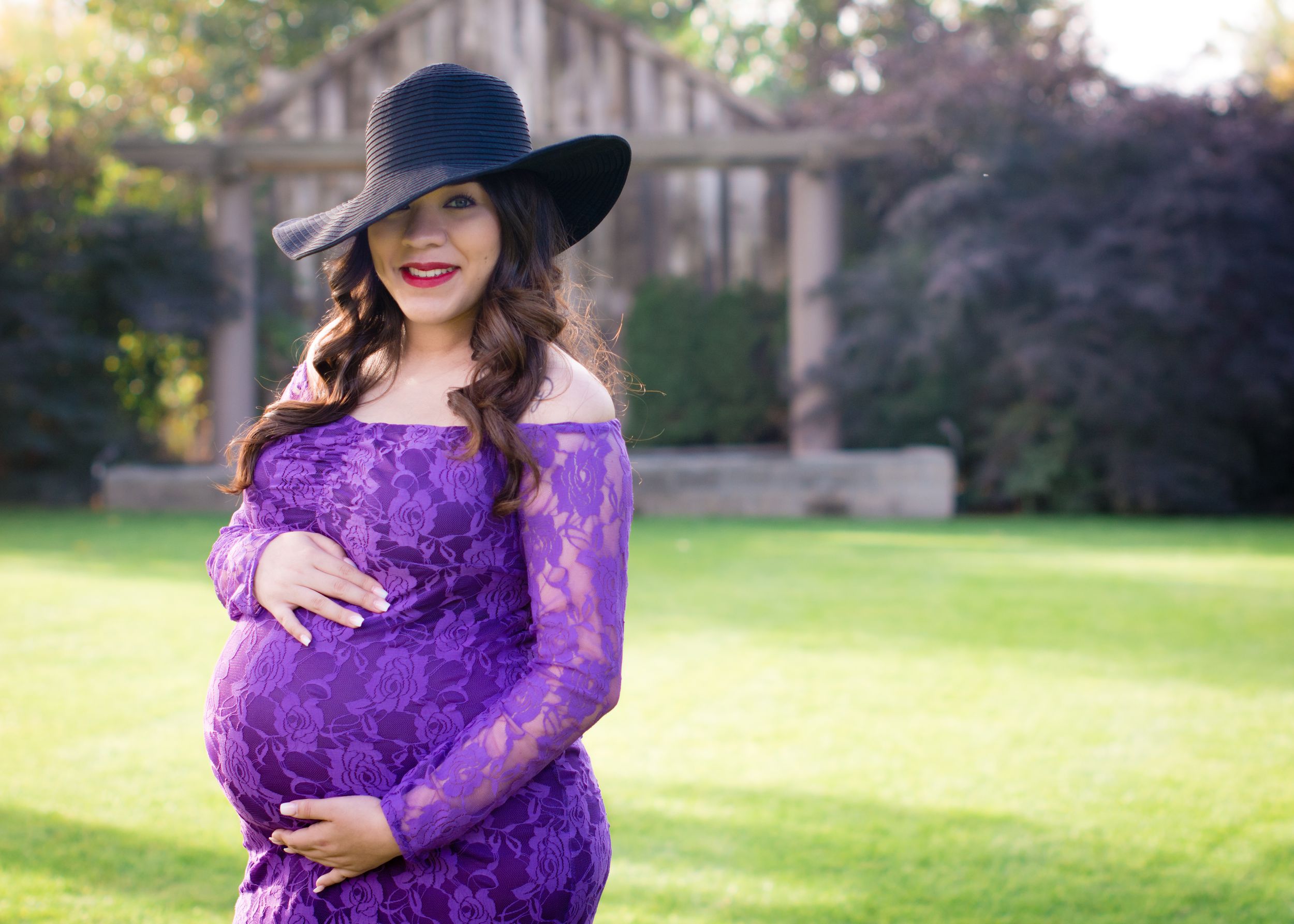 A maternity photo of someone in a purple lace dress and wide-brimmed hat standing in a garden setting.