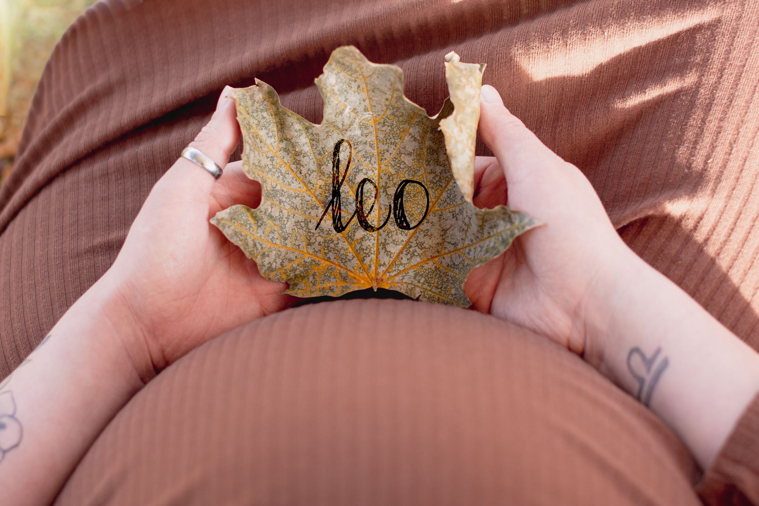 Pregnant woman holding an autumn leaf with the name Leo written on it, showcasing a creative fall maternity photo idea.