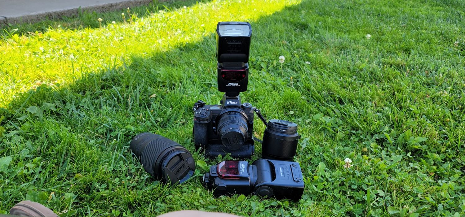 Professional camera equipment including DSLR body, lens and flash unit arranged on green grass in sunlight.