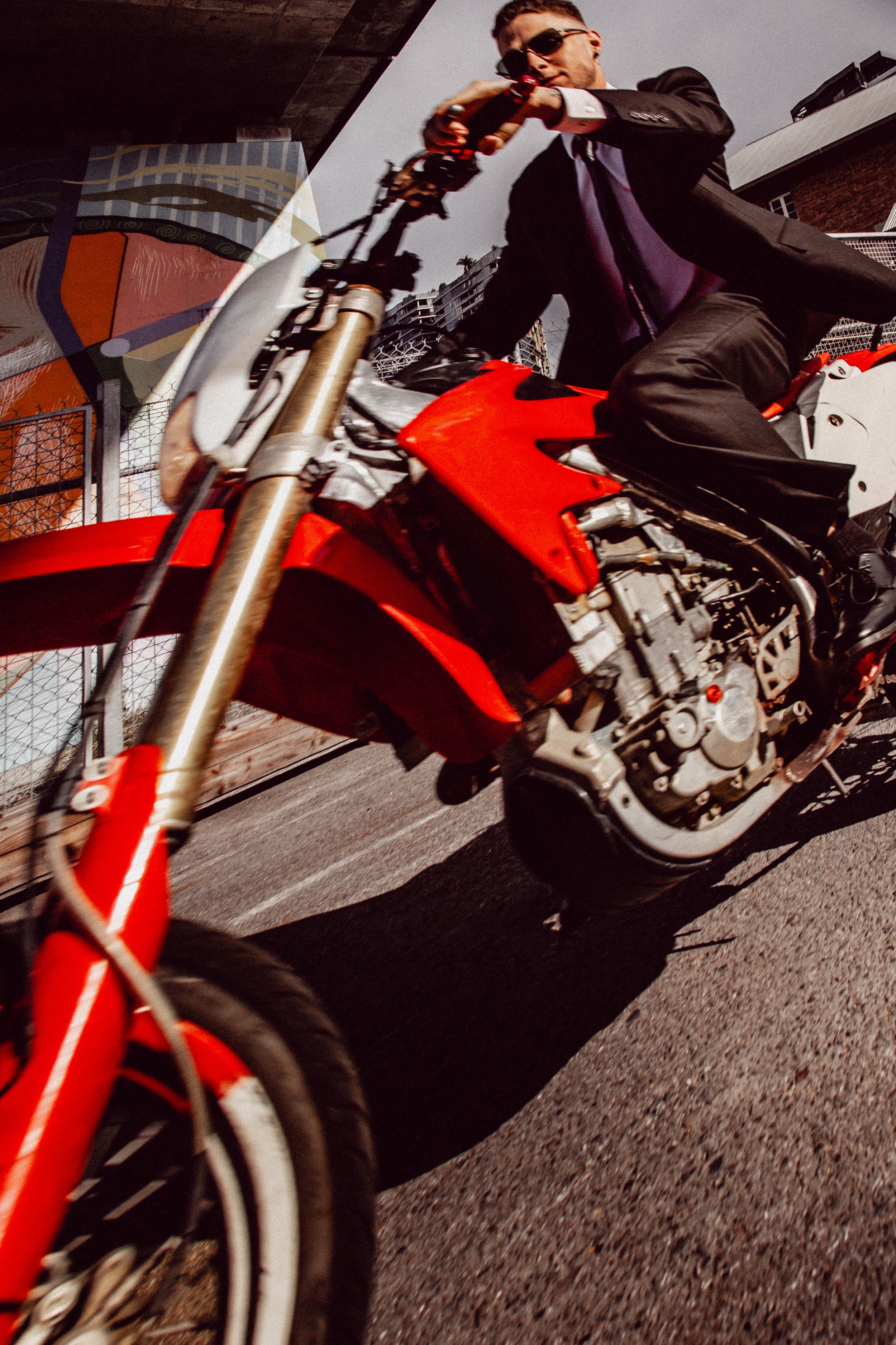 A motorcyclist in a black suit rides a bright red motorbike on a city street with dramatic low angle perspective.