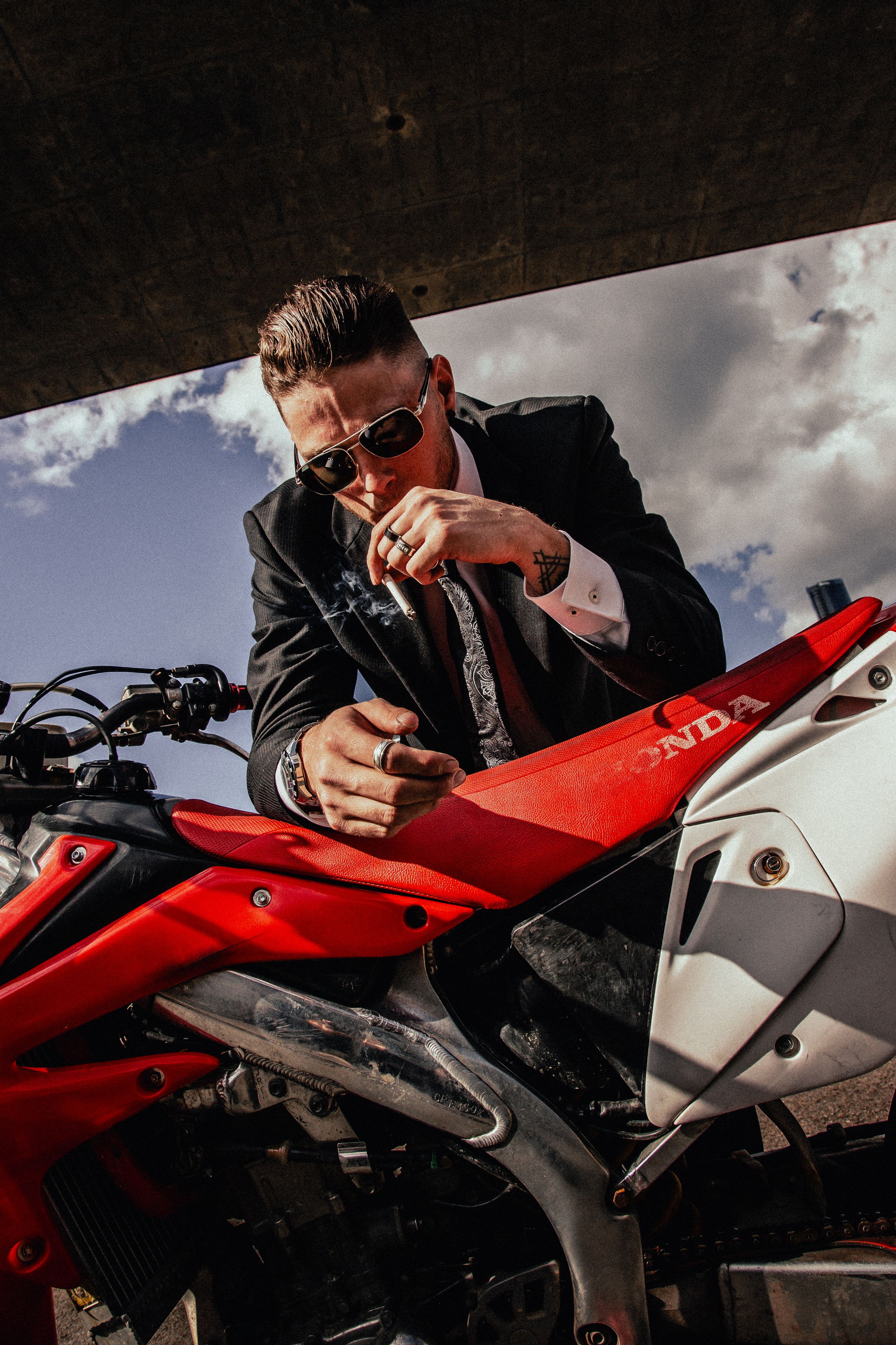 Professional in a black suit and sunglasses checking a red Honda sport motorcycle against a cloudy sky backdrop.