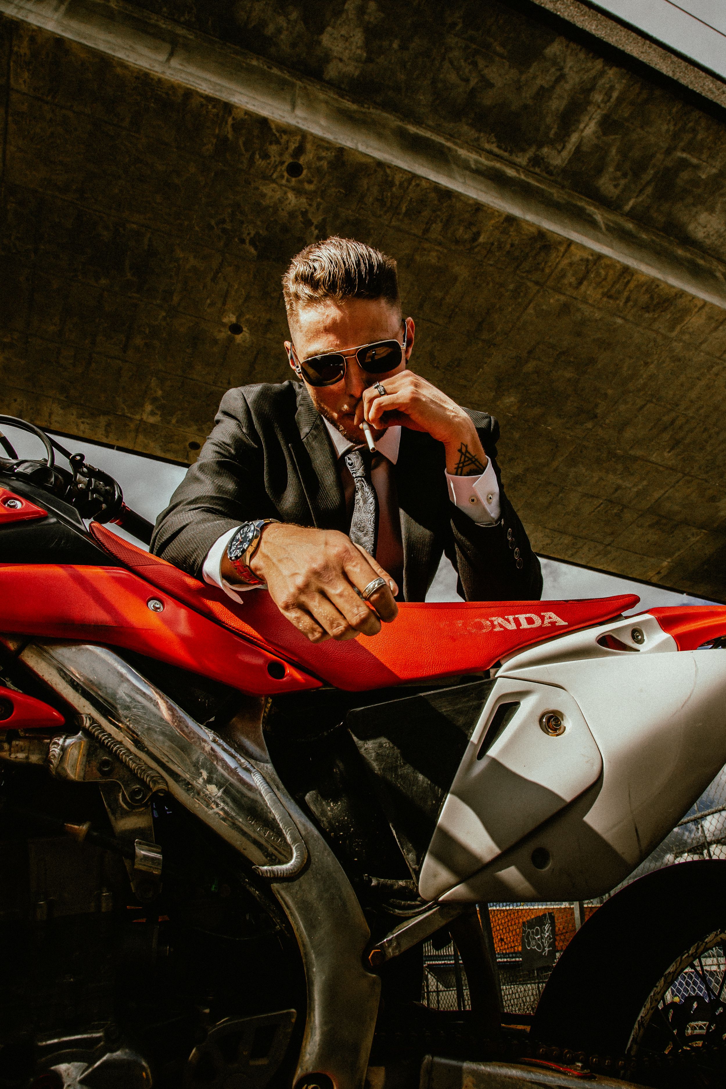 Stylish person in business suit and sunglasses posing coolly next to a red Honda motorcycle under concrete structure.