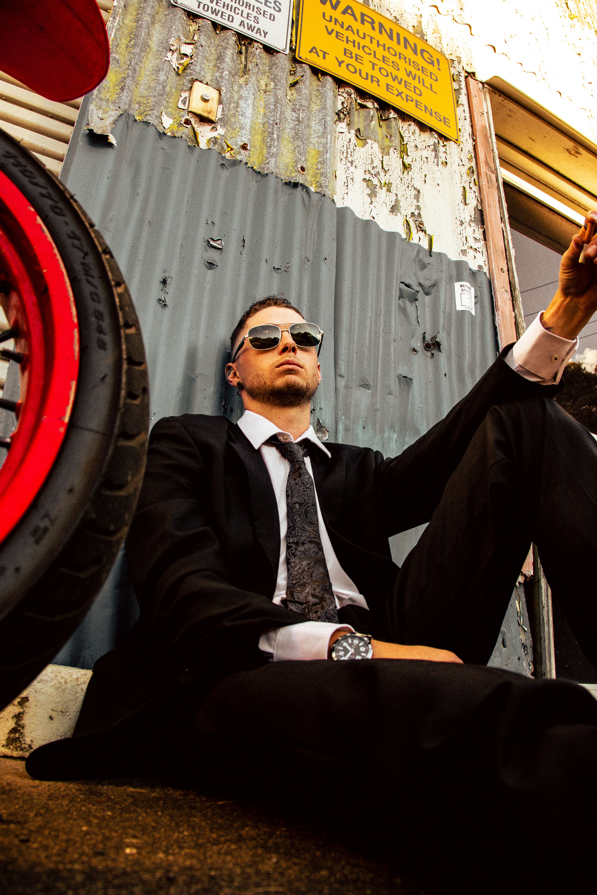 A businessman in a black suit and sunglasses sits casually against an industrial backdrop with corrugated metal walls.