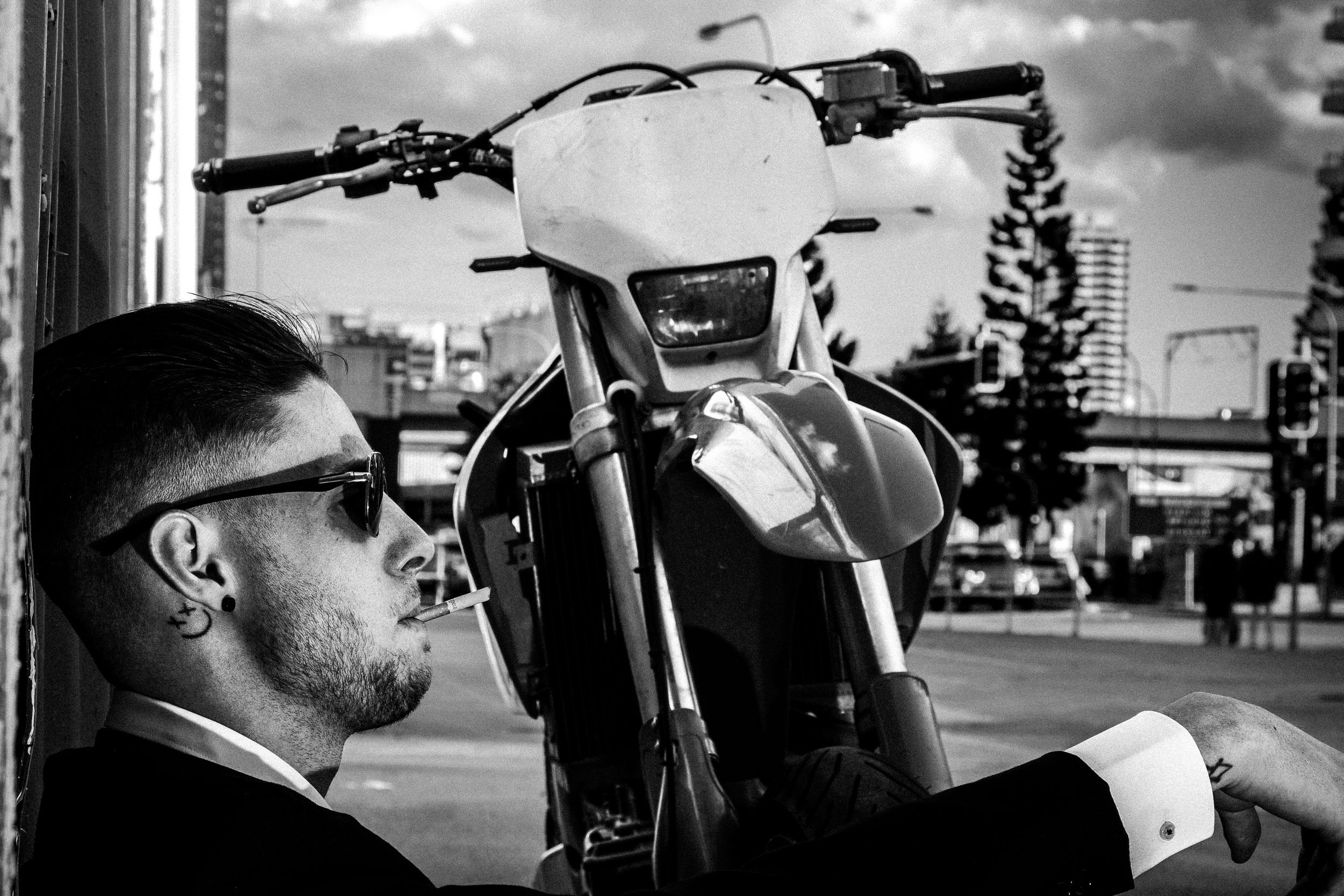 Black and white urban portrait showing a figure in sunglasses next to a motorcycle against a cityscape backdrop.