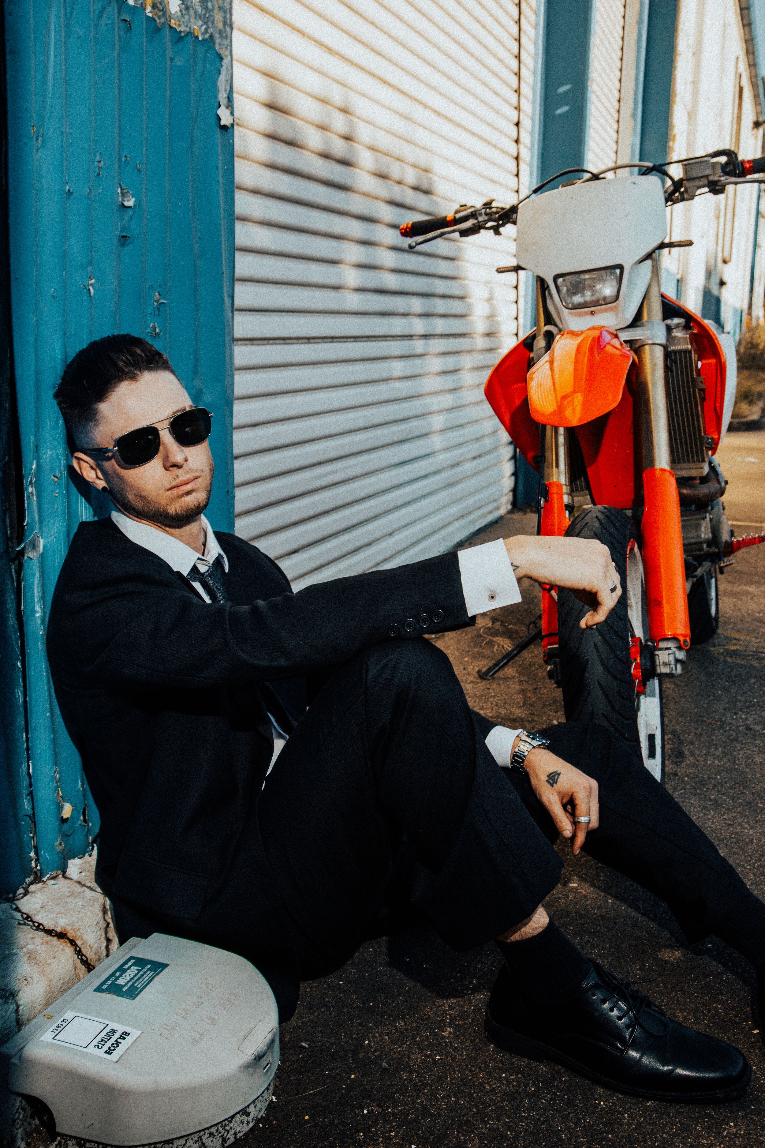 Stylish shot of someone in formal attire sitting beside an orange dirt bike against industrial metal shutters.