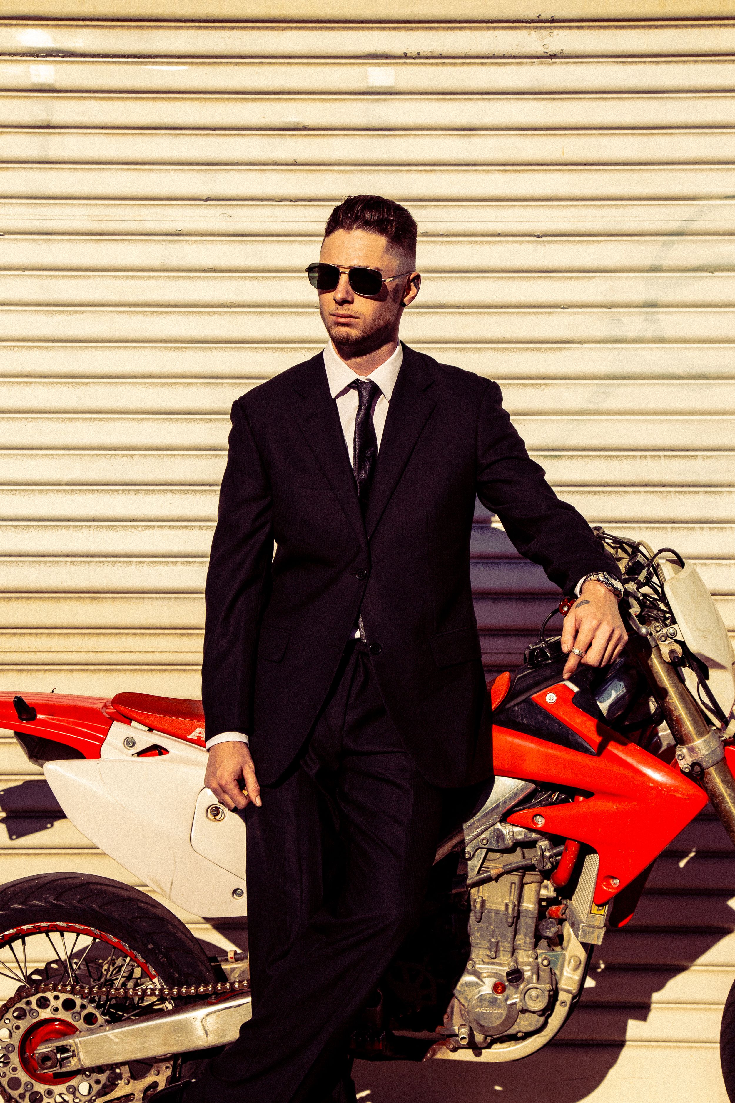 A motorcyclist in a black suit and sunglasses poses with a red and white sport bike against a striped wall background.