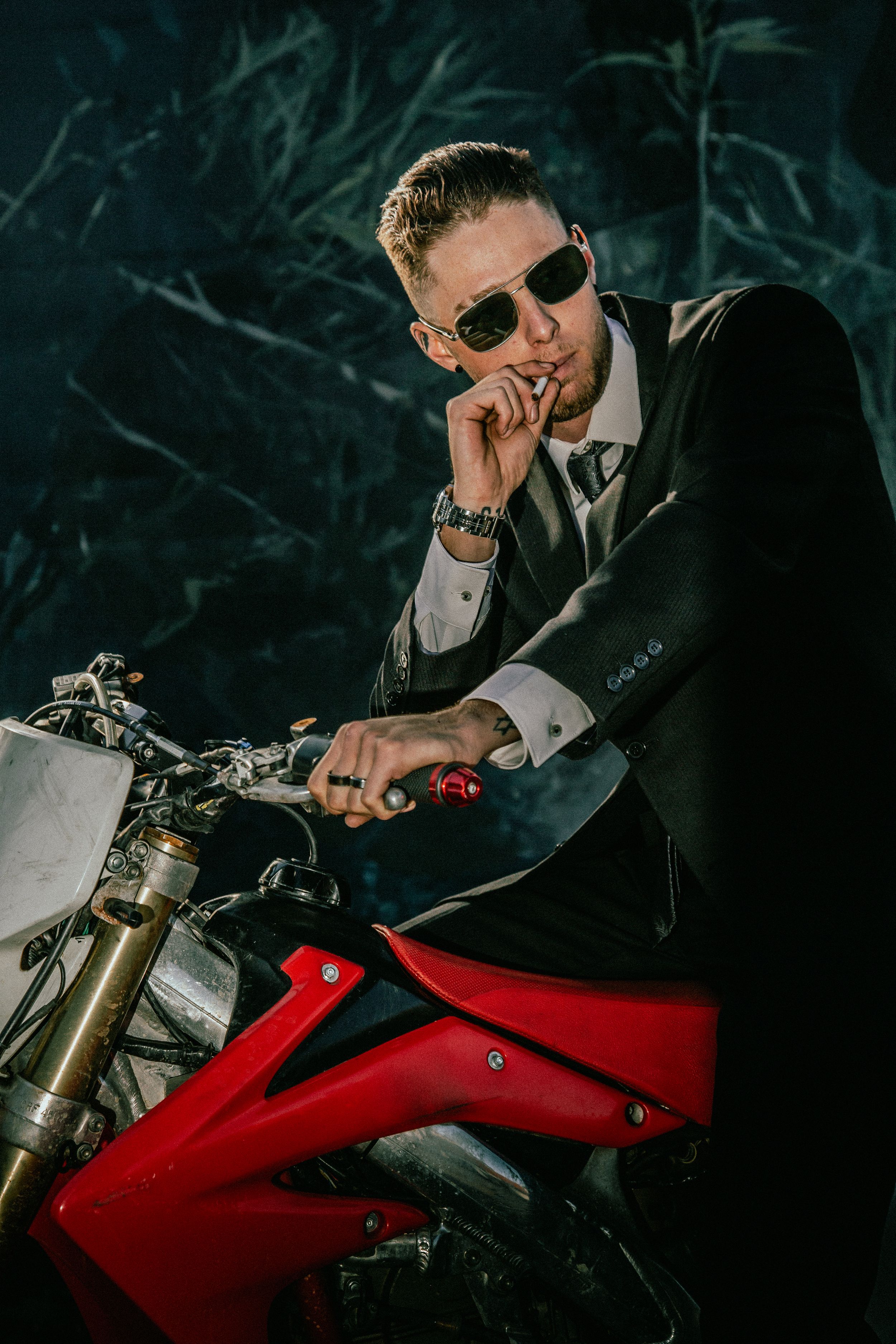 A person in a dark suit and sunglasses sits thoughtfully on a red and silver motorcycle against a moody background.