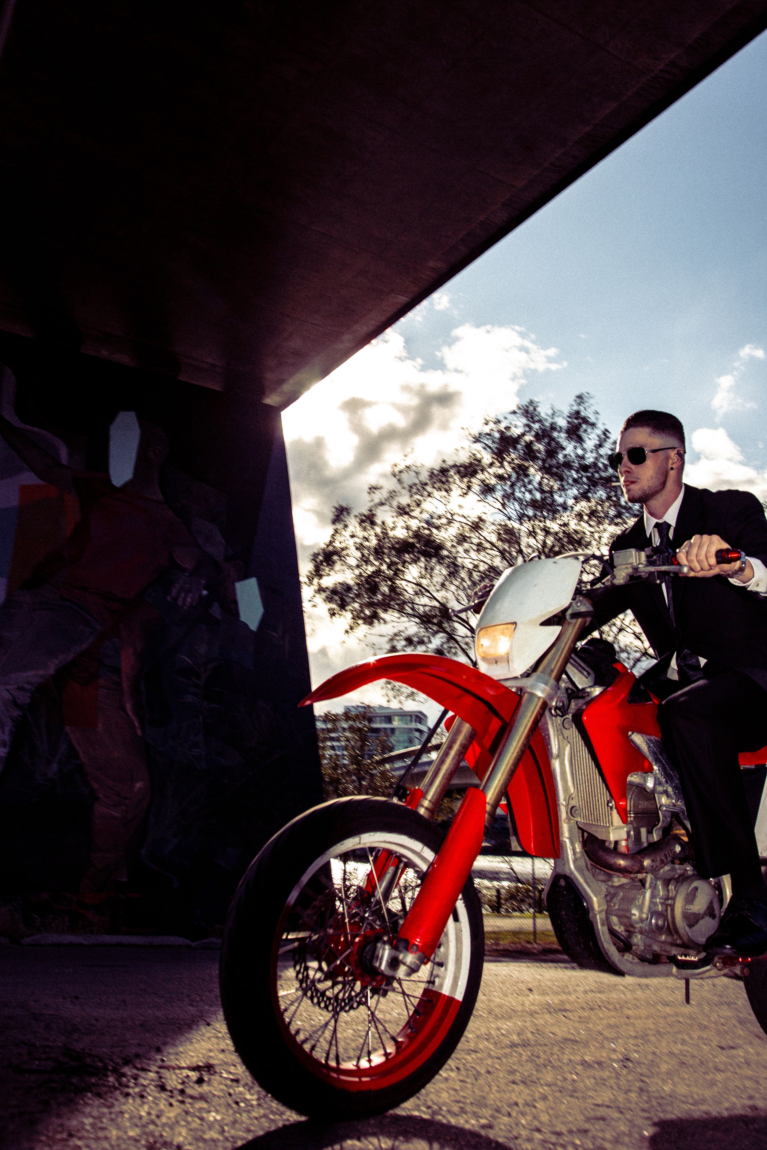 A motorcycle rider in formal attire sits on a red dirt bike under an overpass during sunset.