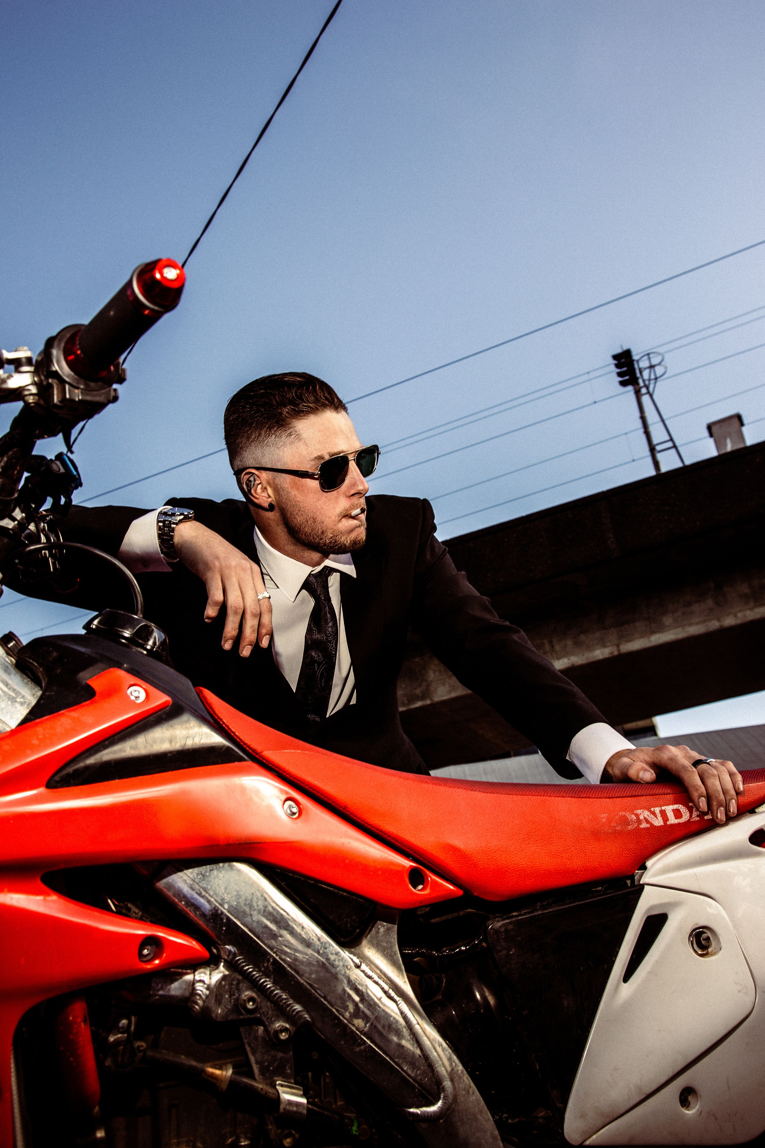 A person in formal attire sits on a red Honda motorcycle against a silhouetted outdoor background.