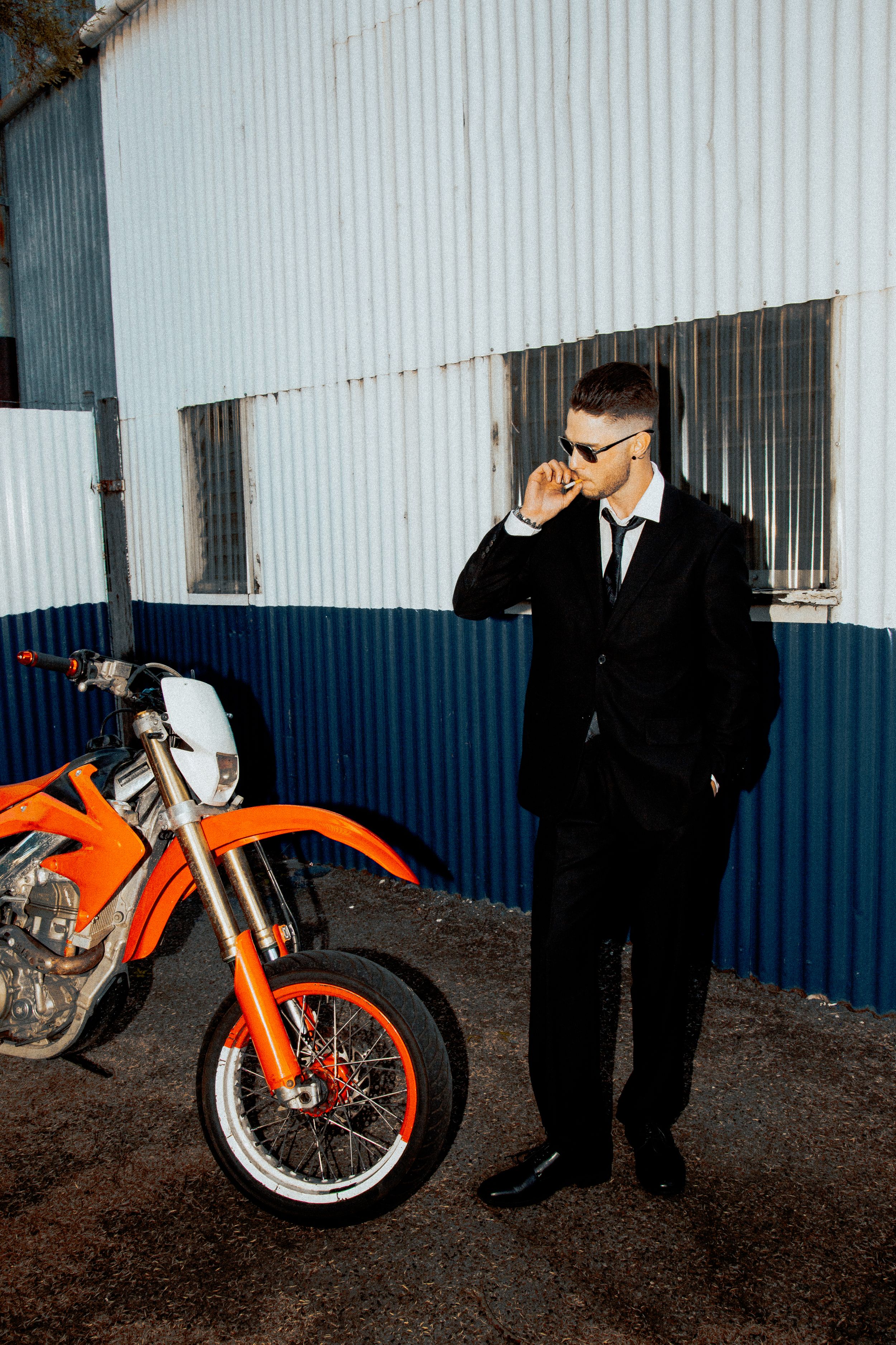 A man in a black suit and sunglasses stands next to an orange dirt bike against a blue and white corrugated wall.