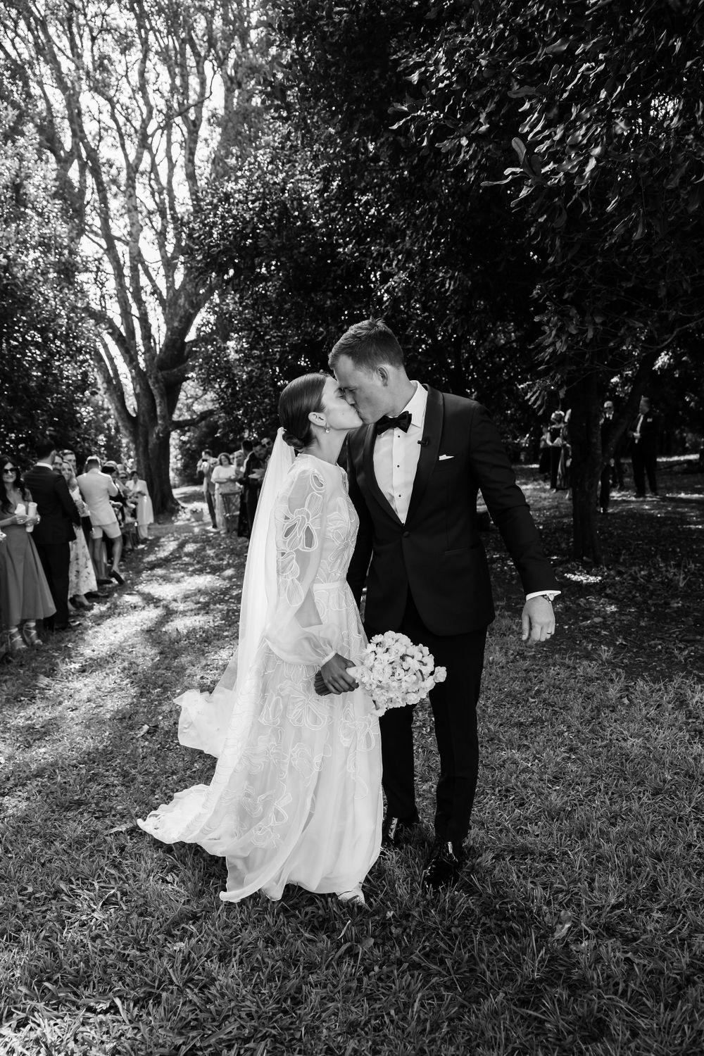 A romantic wedding couple shares a moment in a garden setting with dappled sunlight.