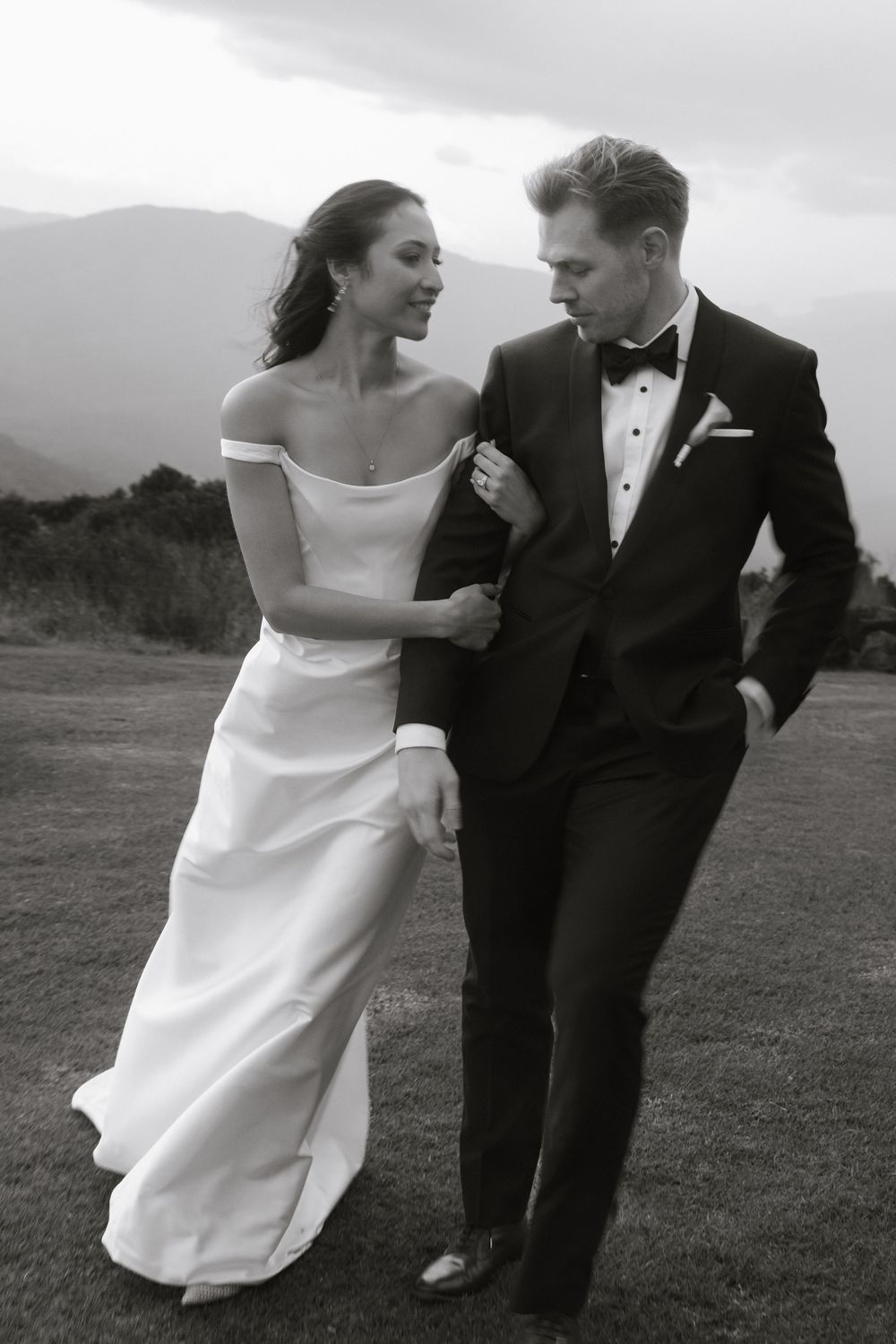 Classic black and white wedding photo of couple walking together outdoors with mountain scenery in the background.